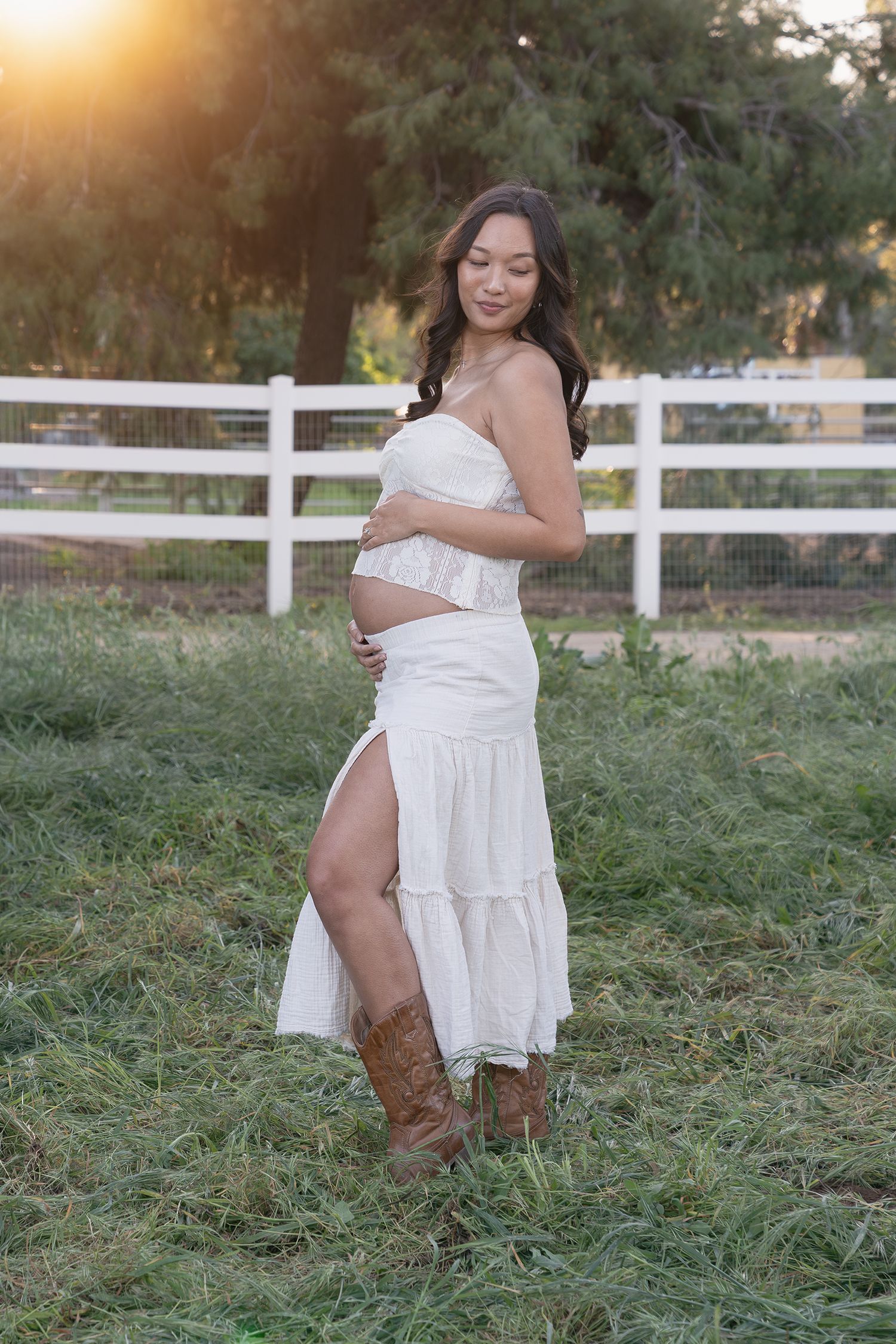 A maternity photo shoot in a rustic setting with cowboy boots and a white dress at sunset by a white fence.