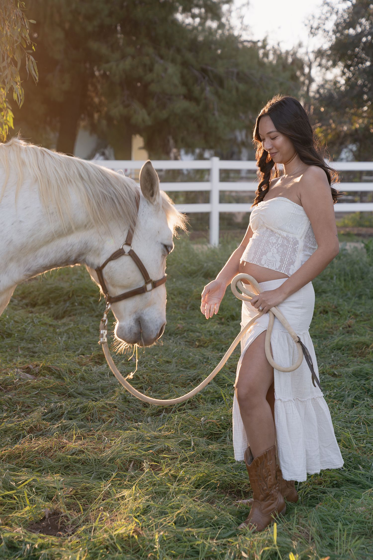 A white horse and woman in a lace dress pose together during golden hour near a white fence.