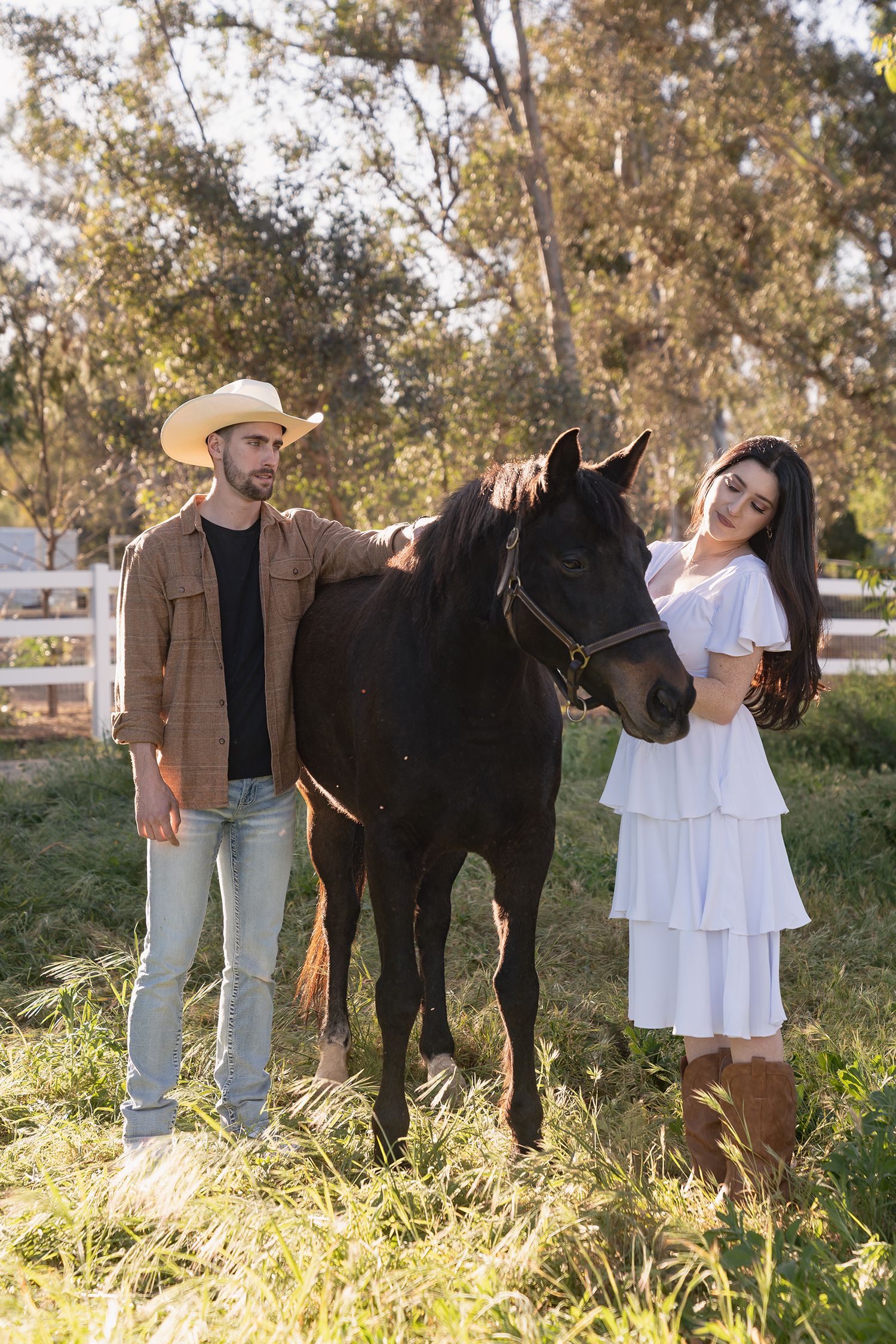 A couple poses with their black horse in a sunny field with white fencing and trees in the background.