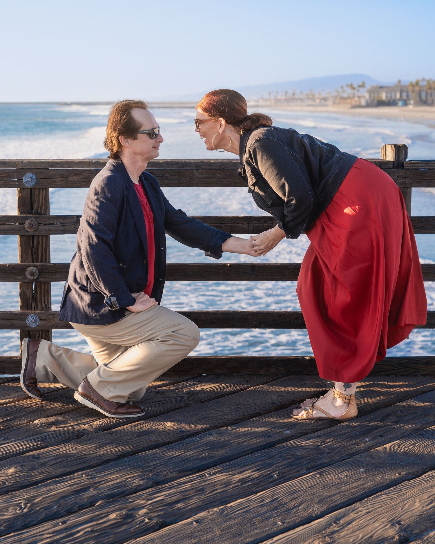A couple sits together on a weathered wooden pier with ocean waves visible below.