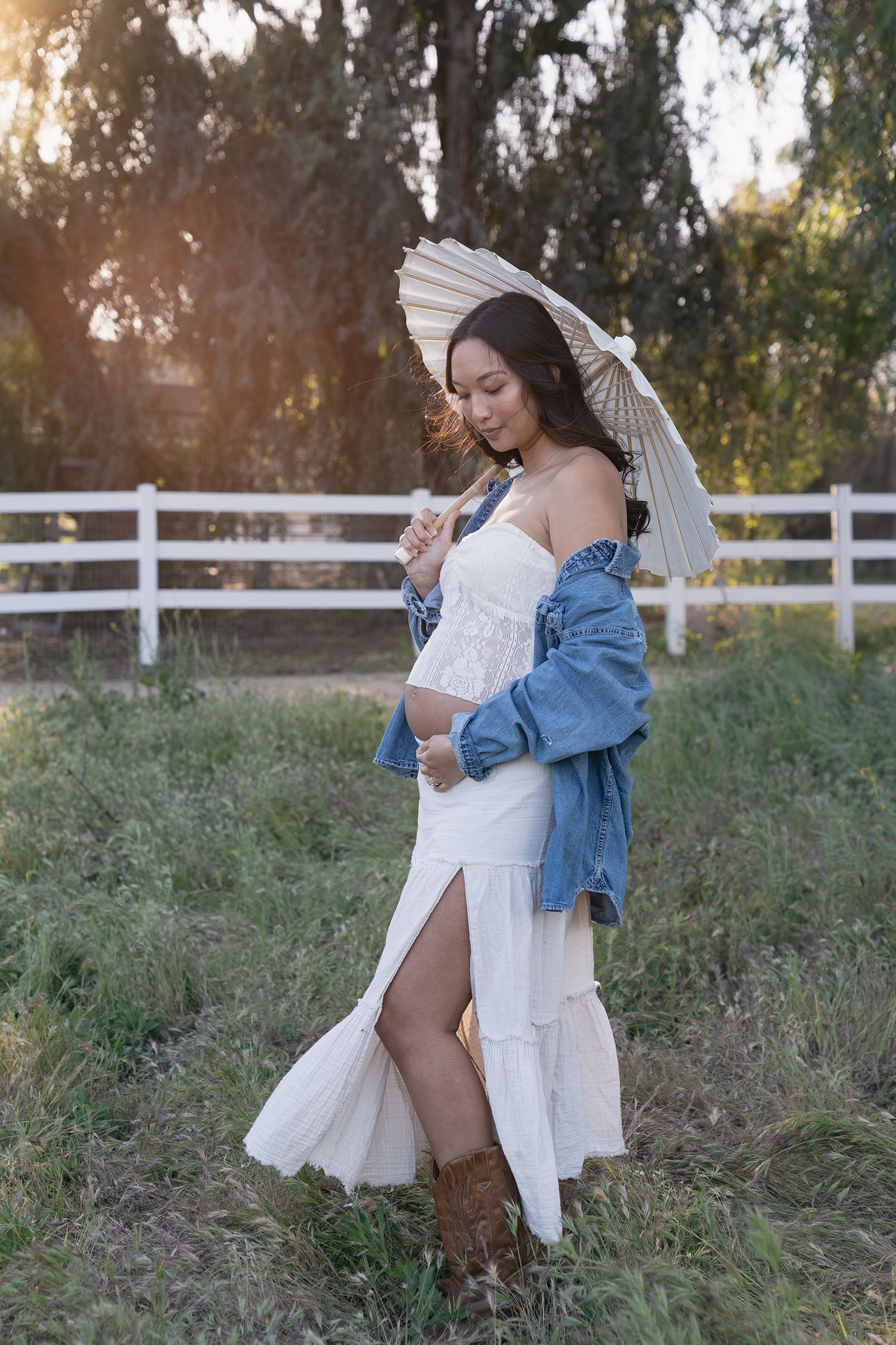 A figure in a white dress and denim jacket poses gracefully against a rustic fence and natural outdoor setting.