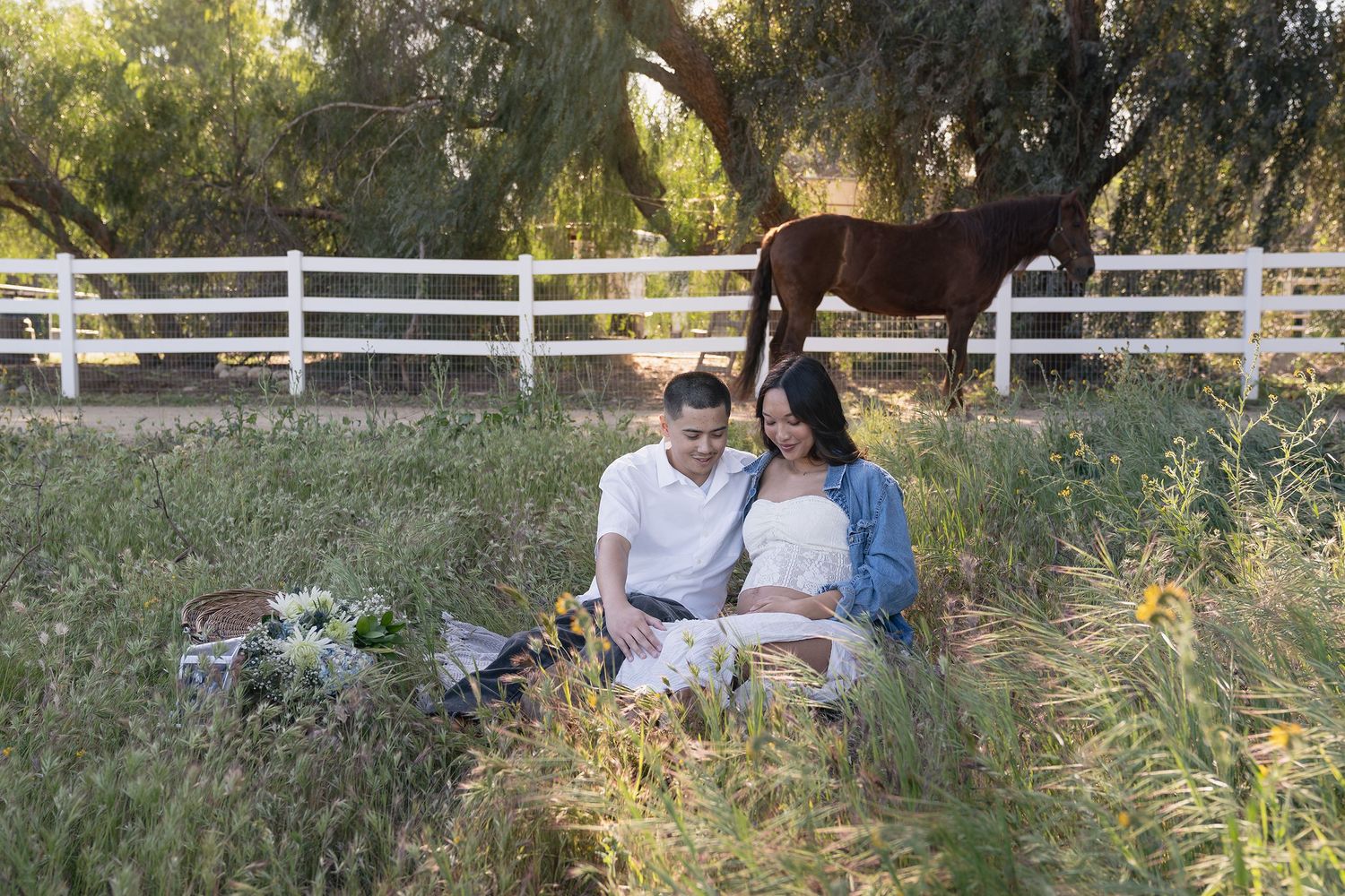 A couple sits together on a blanket having a romantic picnic in a sunlit meadow with tall grass and trees.