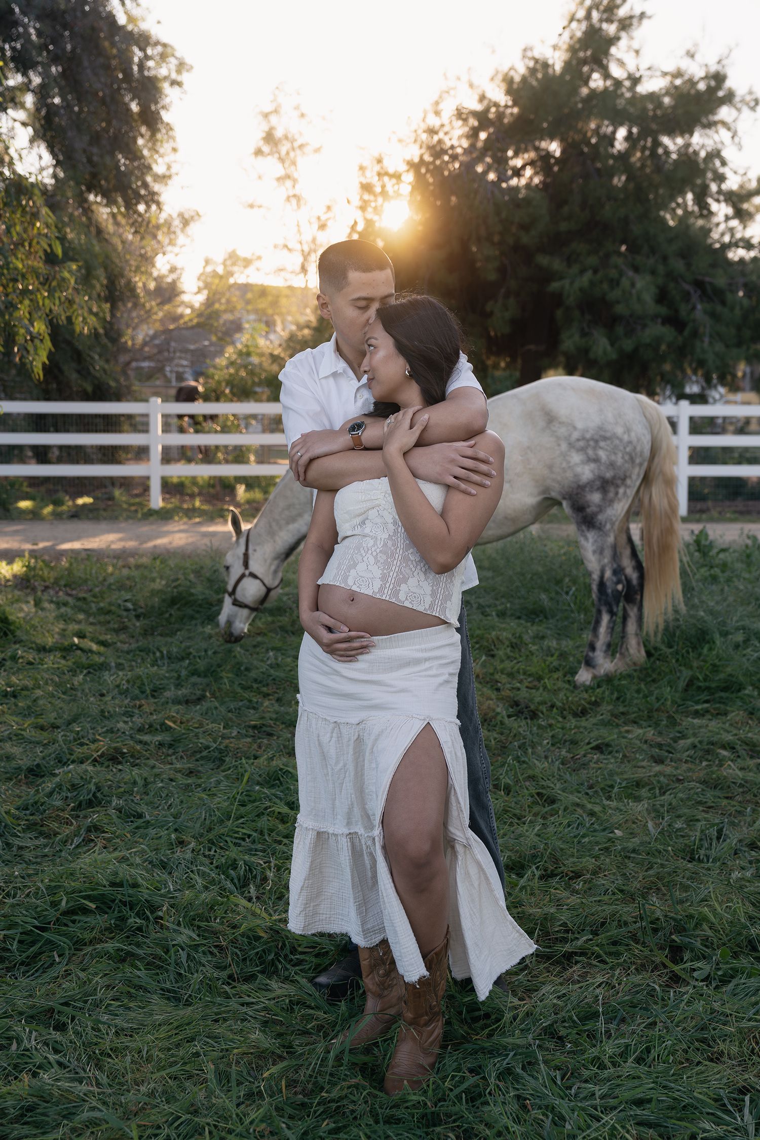 Mother and child share tender moments outdoors at sunset with white horse in rustic farm setting.