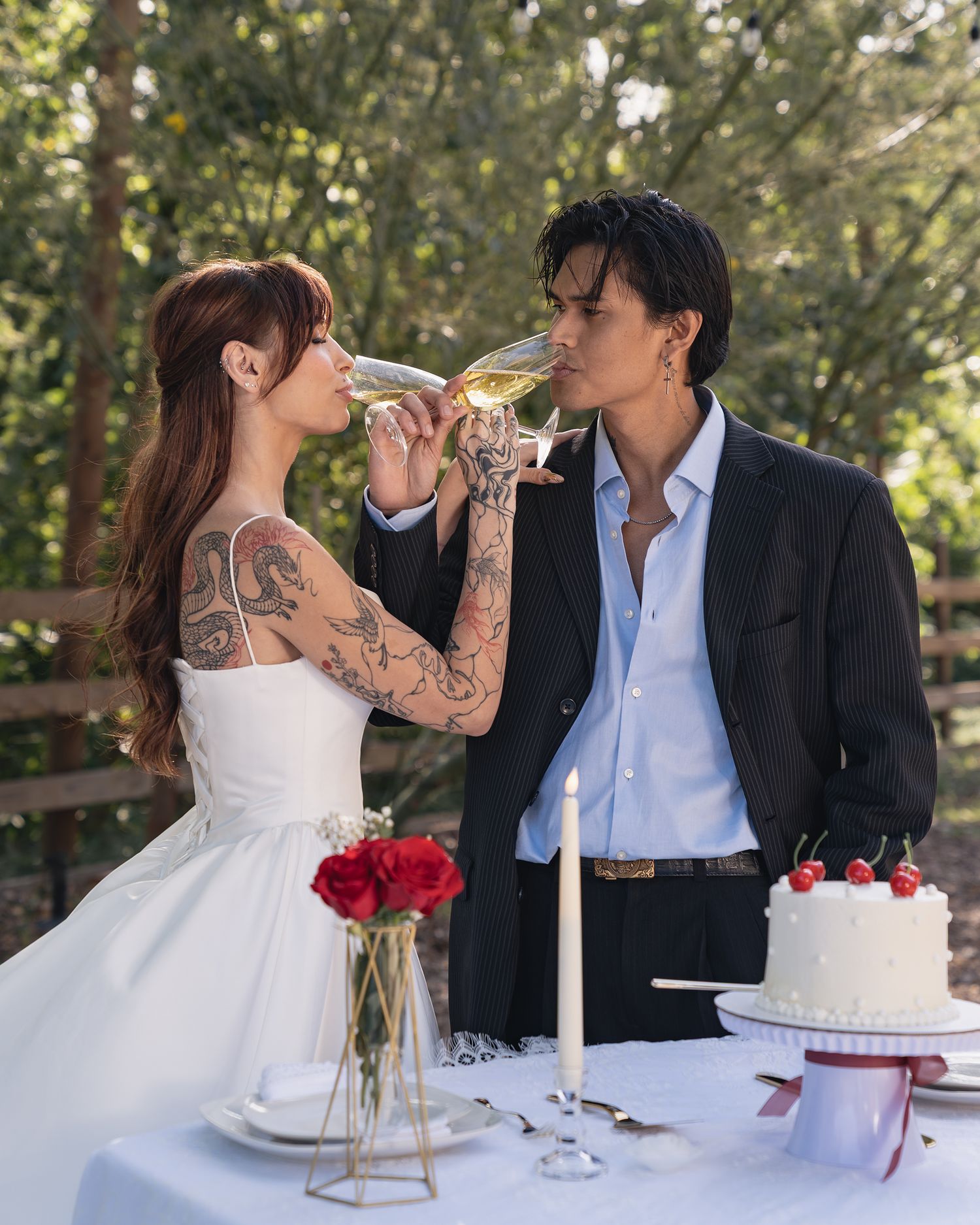 Tattooed bride in white dress sips champagne with groom in black suit at outdoor wedding table with cake.