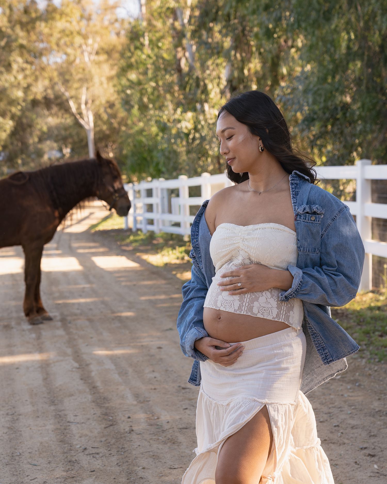A woman in a white dress and denim jacket poses with a brown horse at a ranch with white fencing in the background.