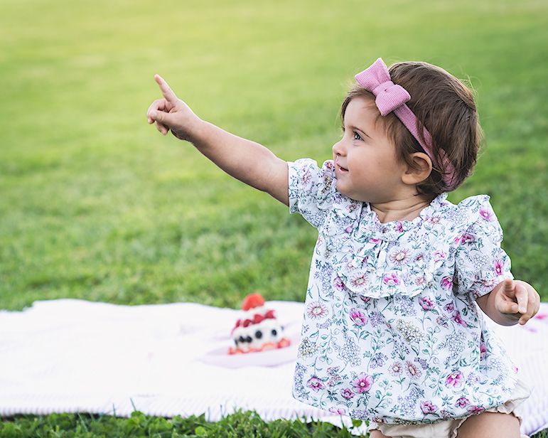 A small child in floral dress points excitedly while sitting on a picnic blanket.