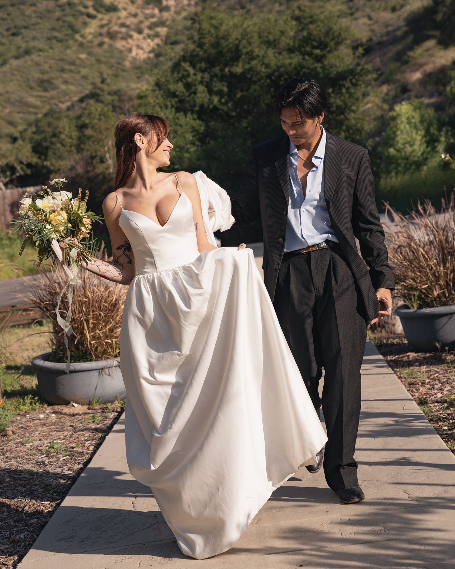 A bride in a white ballgown holds flowers while walking with a suited groom on a garden path with green hills behind them.