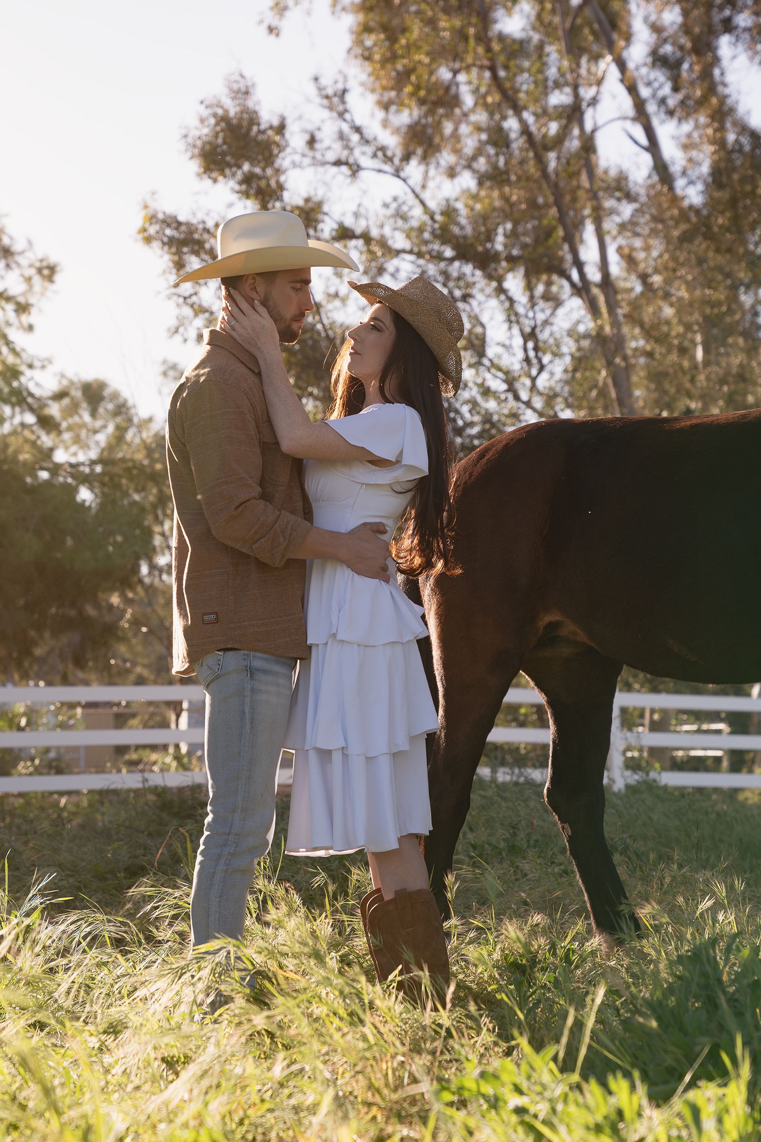 A couple embraces in a romantic moment at sunset on a ranch with white fencing and tall grass in the background.
