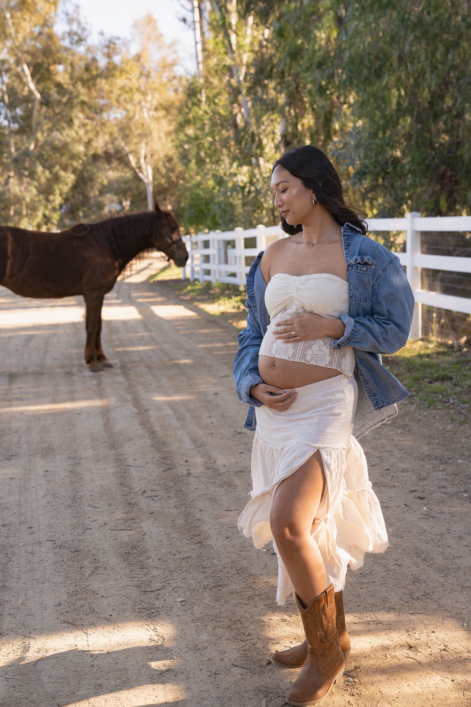 A couple in casual attire embraces and dances together on a dirt path with white fencing and trees in the background.