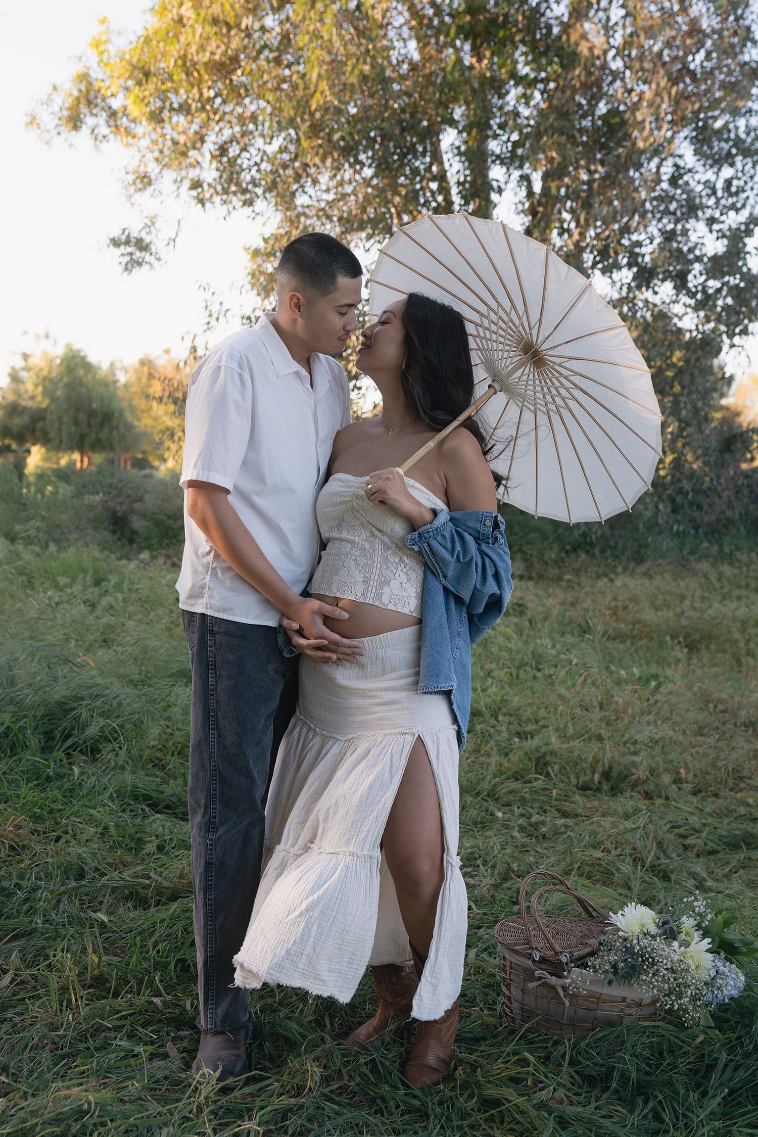Romantic outdoor photoshoot with white parasols and elegant white dress against a natural grassy backdrop at sunset.
