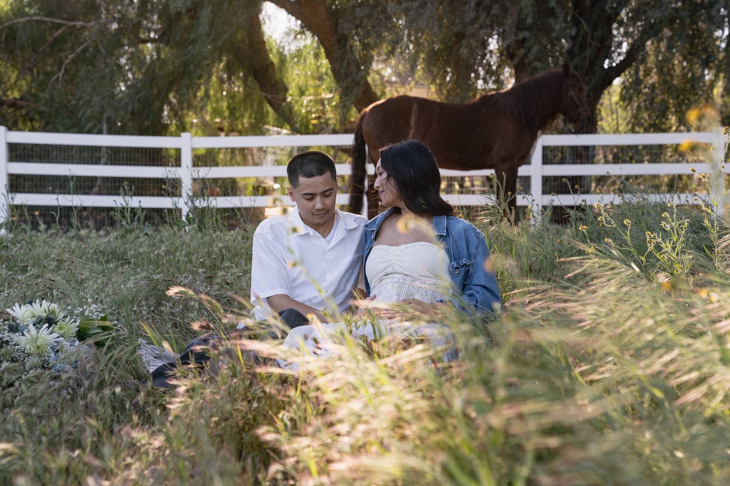 A couple enjoys a romantic picnic near a white fence with a horse grazing in the background.