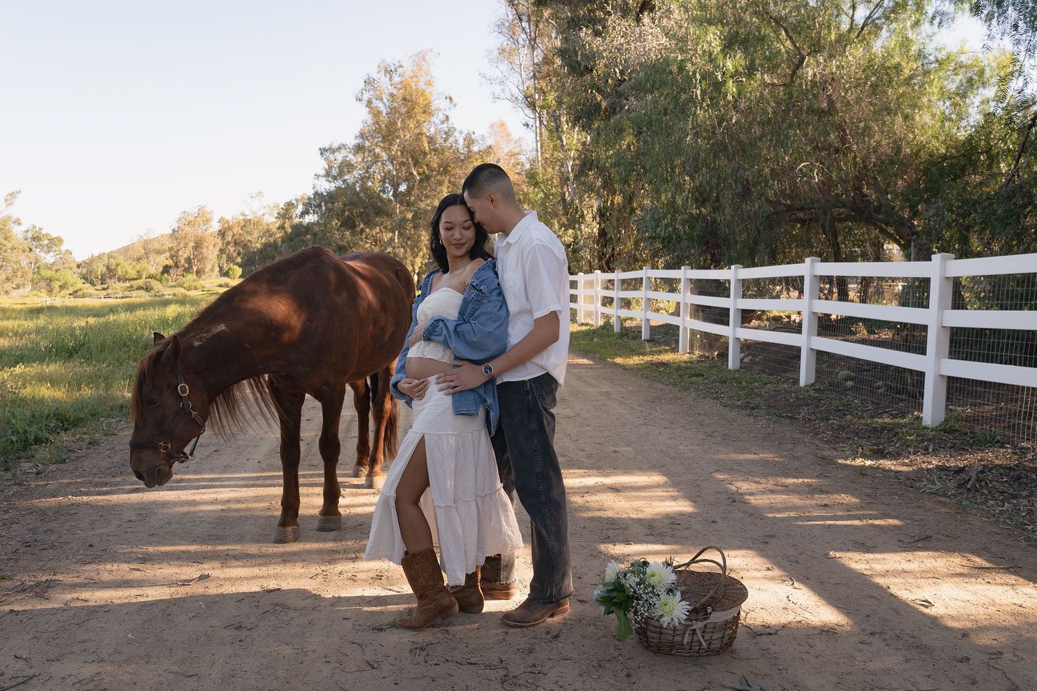A couple poses with their horse near a white fence at sunset during an outdoor photo session.