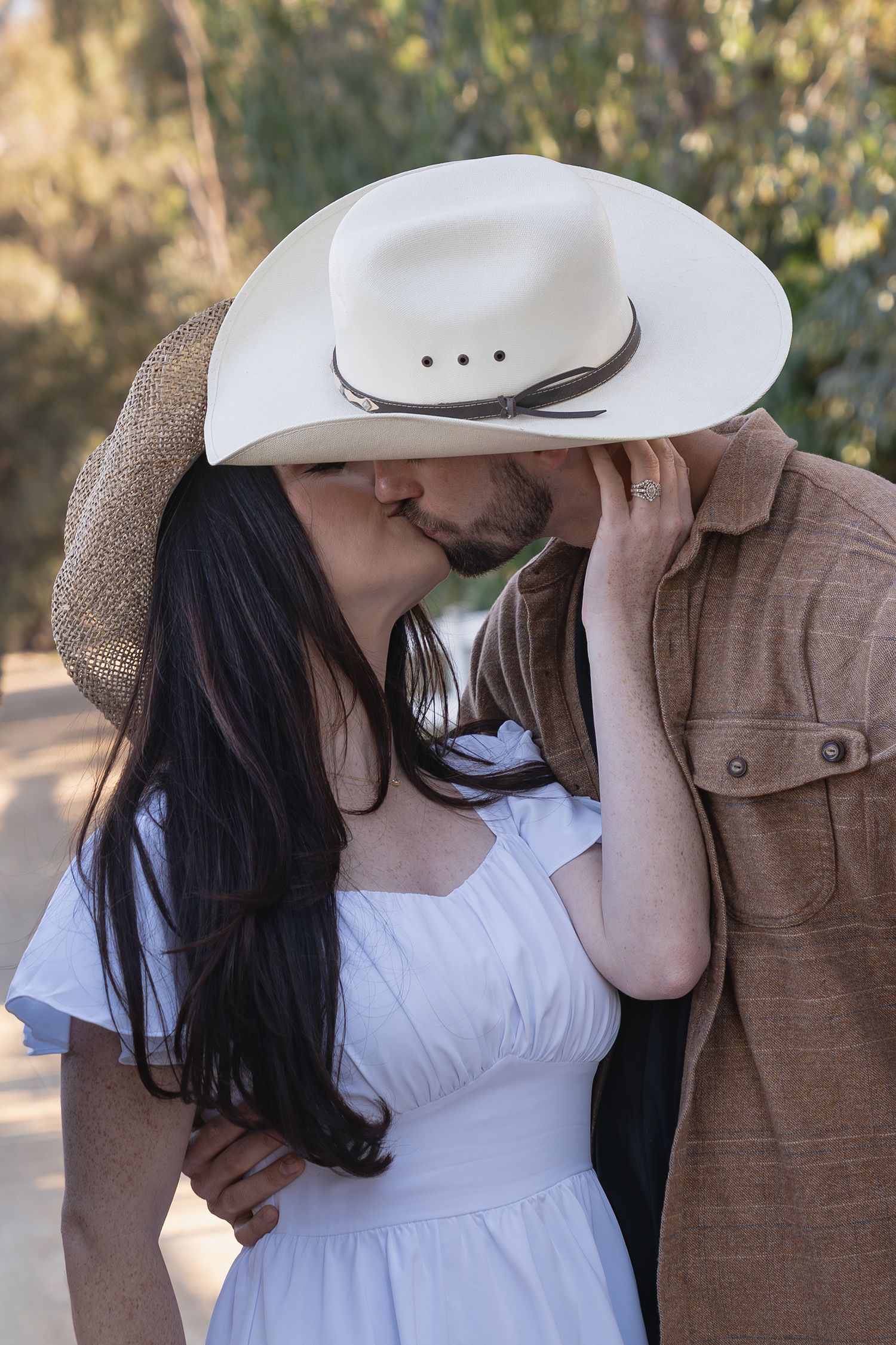 A couple in western attire shares an intimate moment, with a white cowboy hat and brown jacket visible in sunlight.
