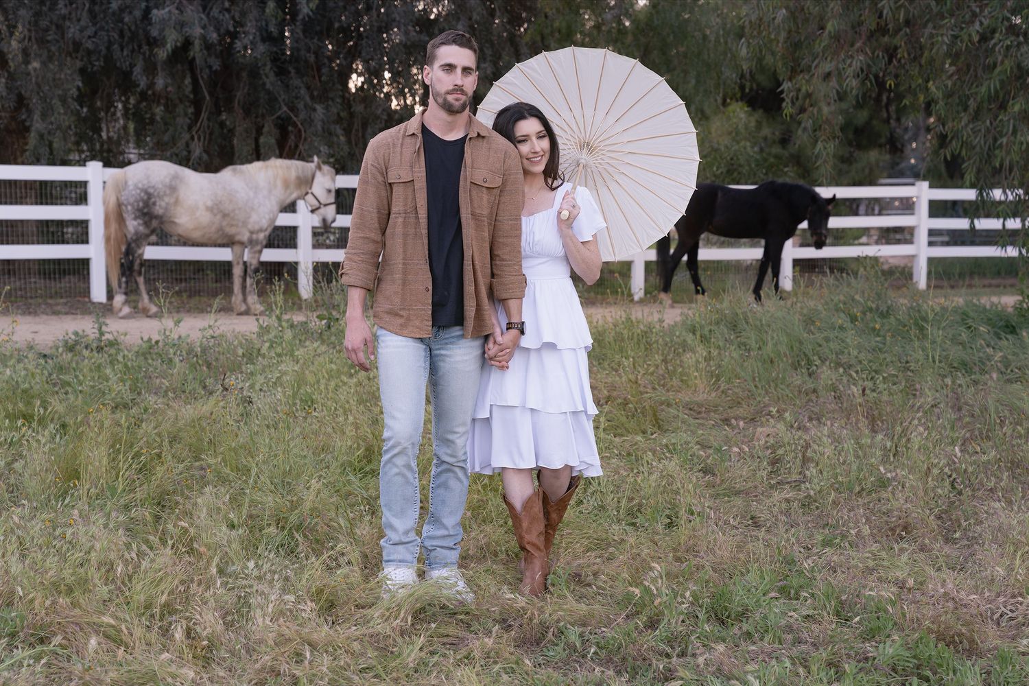 A couple poses together in a grassy field with white parasols and rustic fencing in the background.