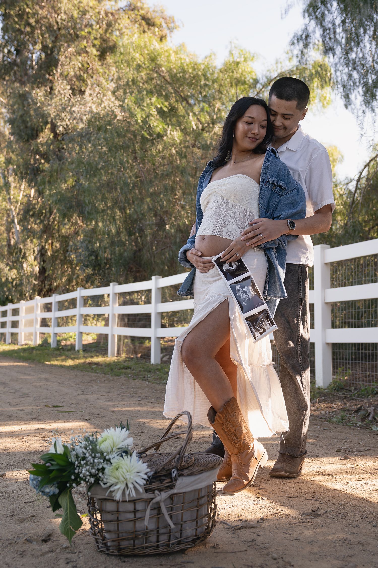 A couple shares an intimate moment together on a rustic farm path with white fencing and trees in the background.