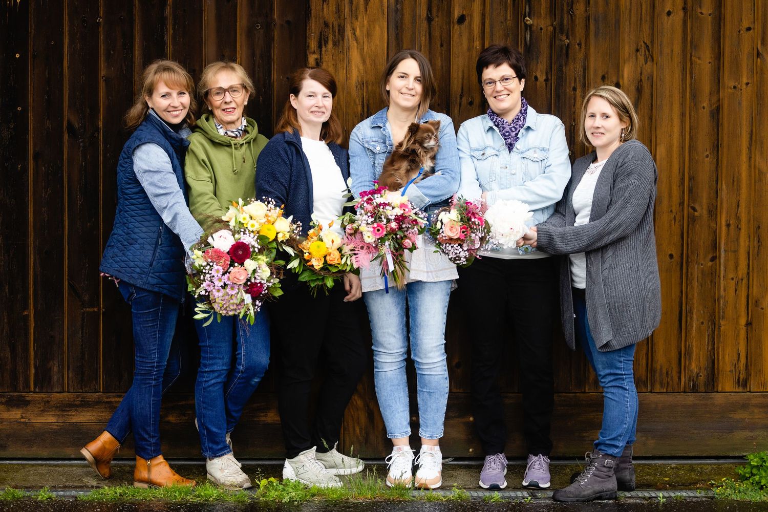 A group stands together holding colorful flower bouquets against a rustic wooden wall background.