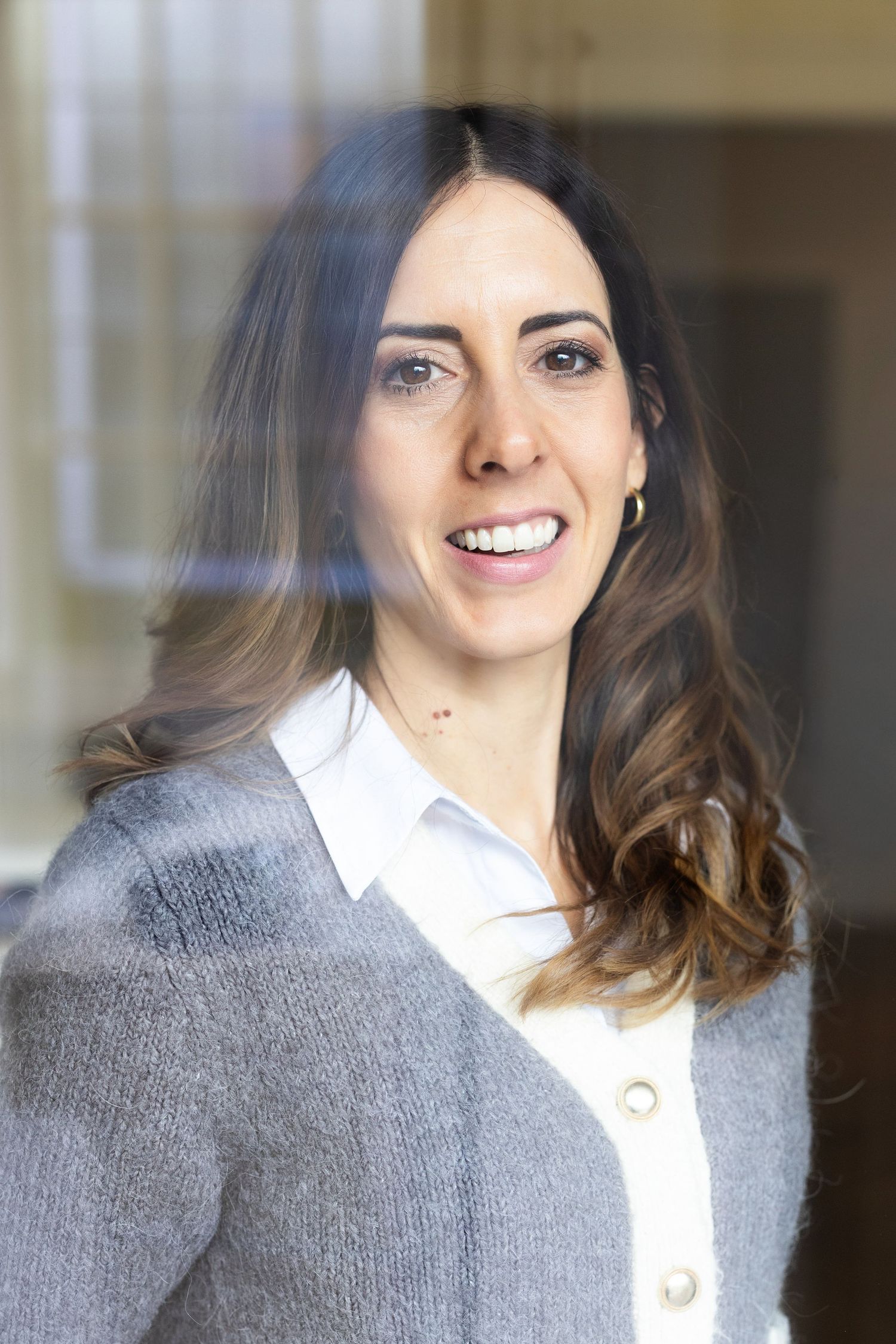 Professional portrait of person in gray cardigan and white blouse smiling at camera in office setting.