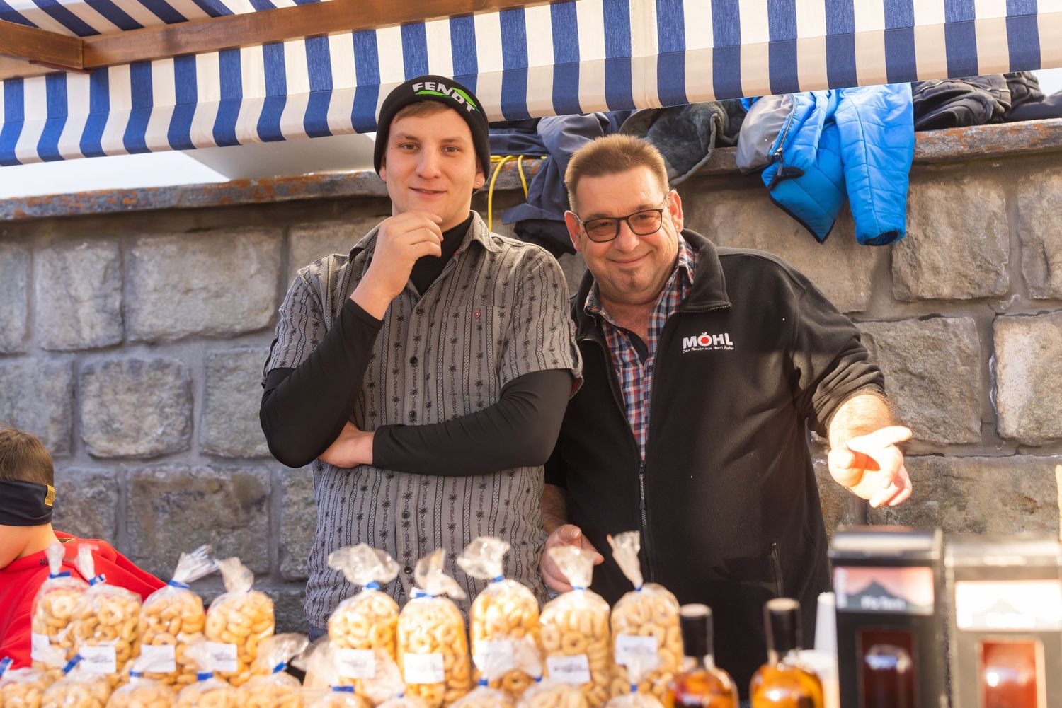 Two people behind market stall with baked goods and blue striped awning overhead.