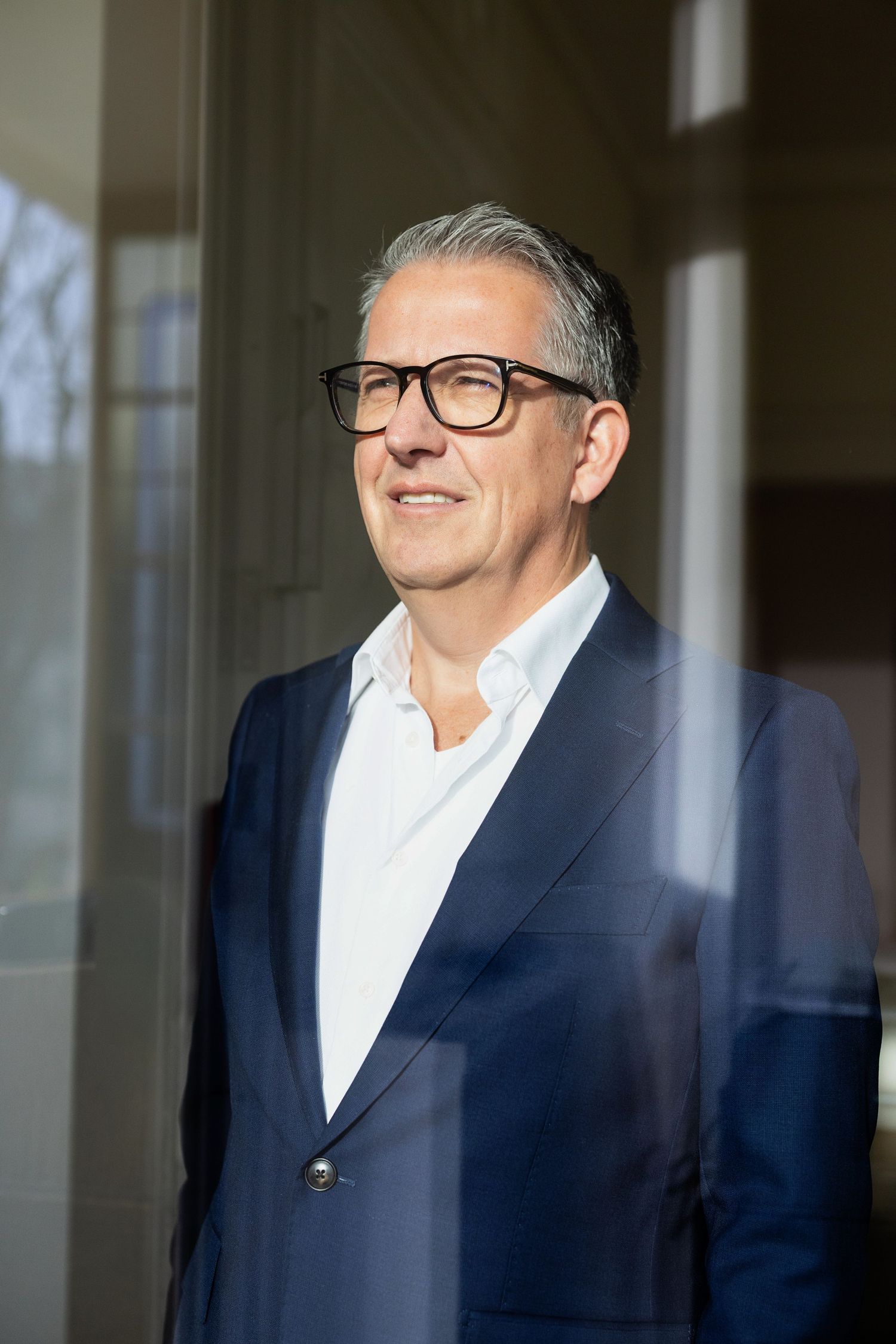 Professional headshot of person in navy suit and glasses standing by window.