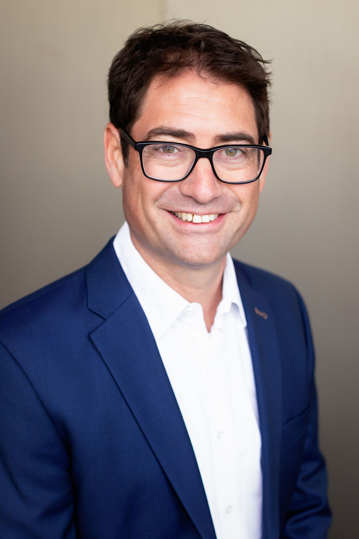 Professional headshots in a navy blazer and white shirt with glasses against a neutral backdrop.
