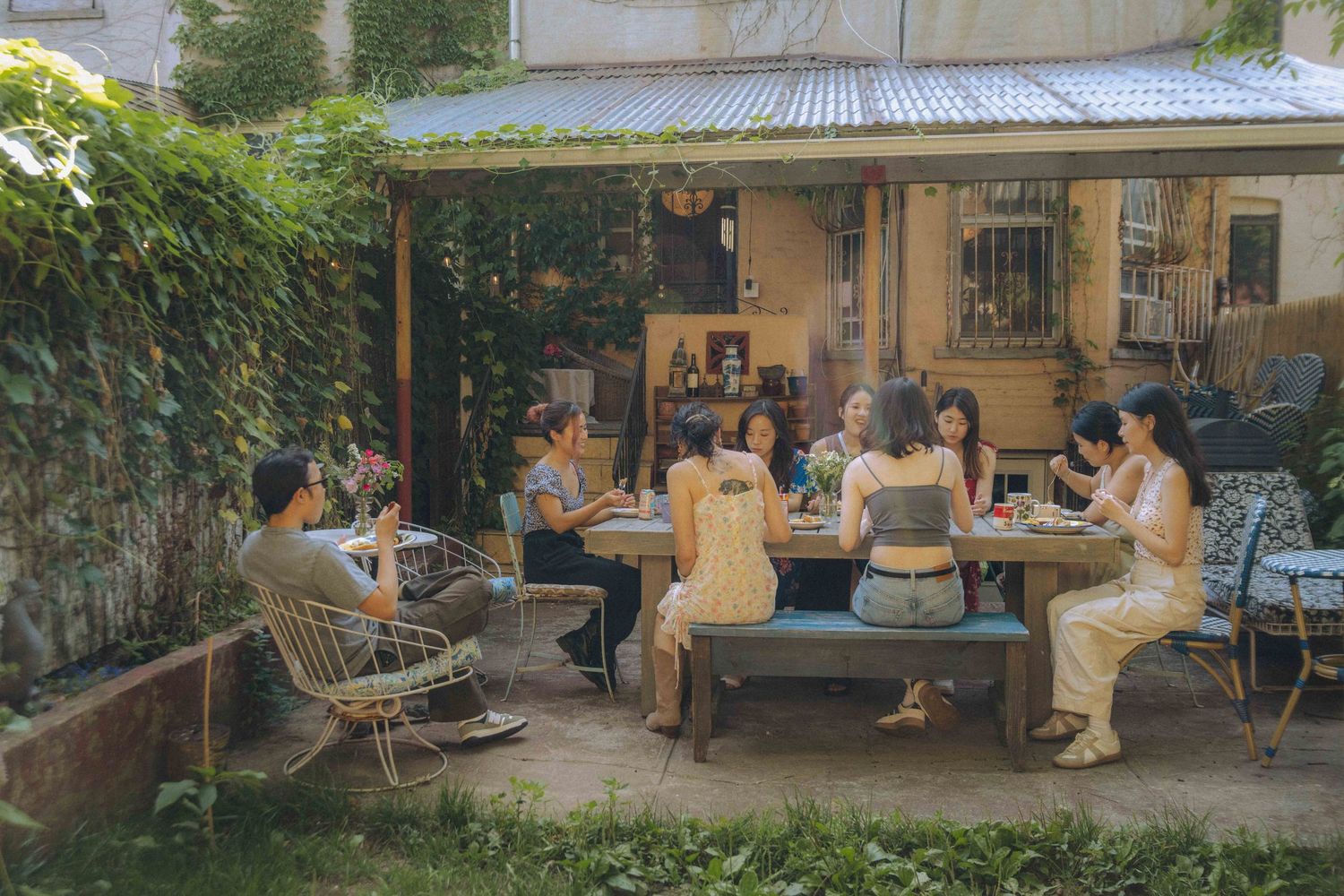 Wide outdoor photo of a small group seated around a long table in a leafy backyard patio, gathered for a meal in the afternoon light.
