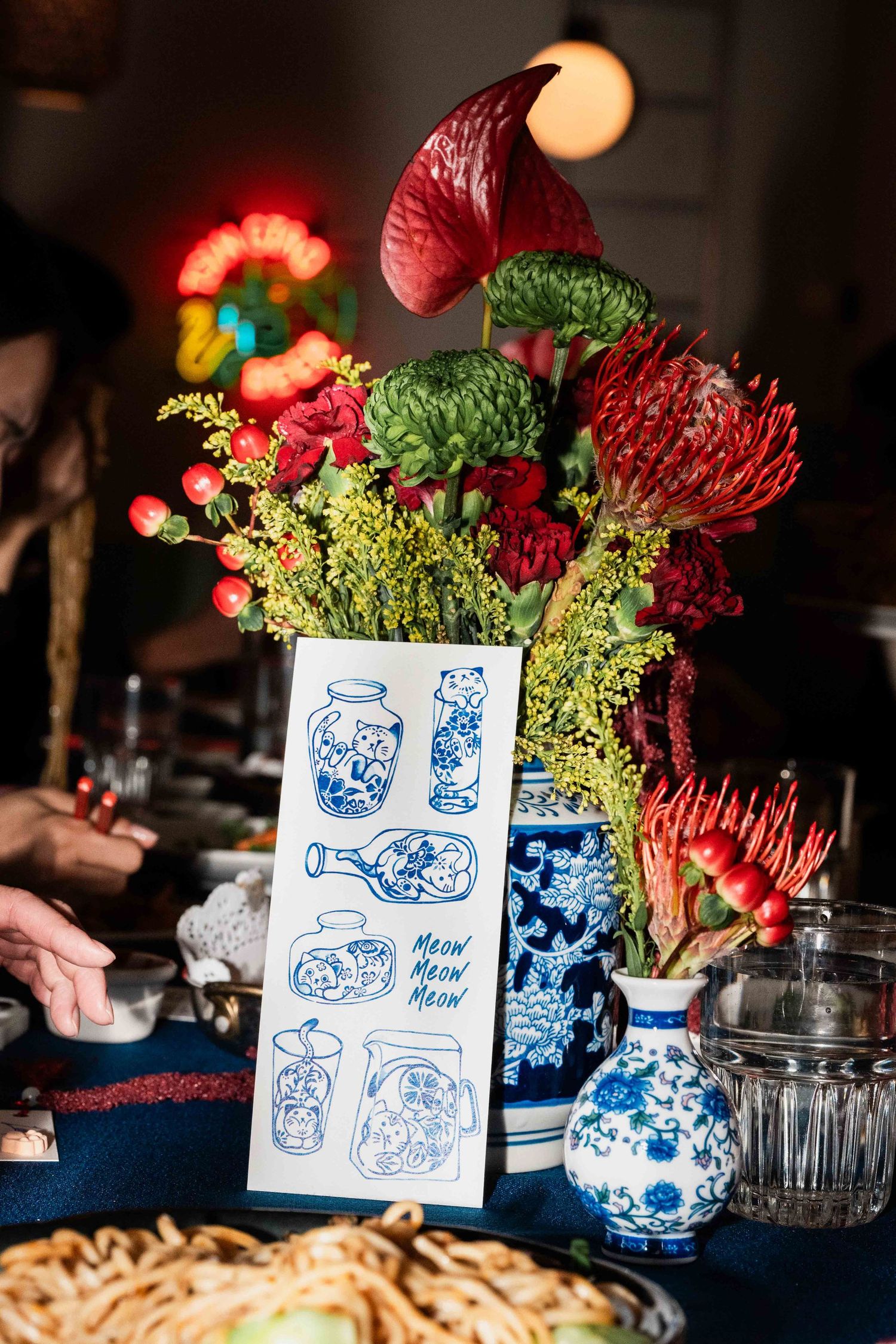 Close-up of a floral centerpiece with red and green blooms beside a blue-and-white illustrated menu card on a dining table, with soft indoor