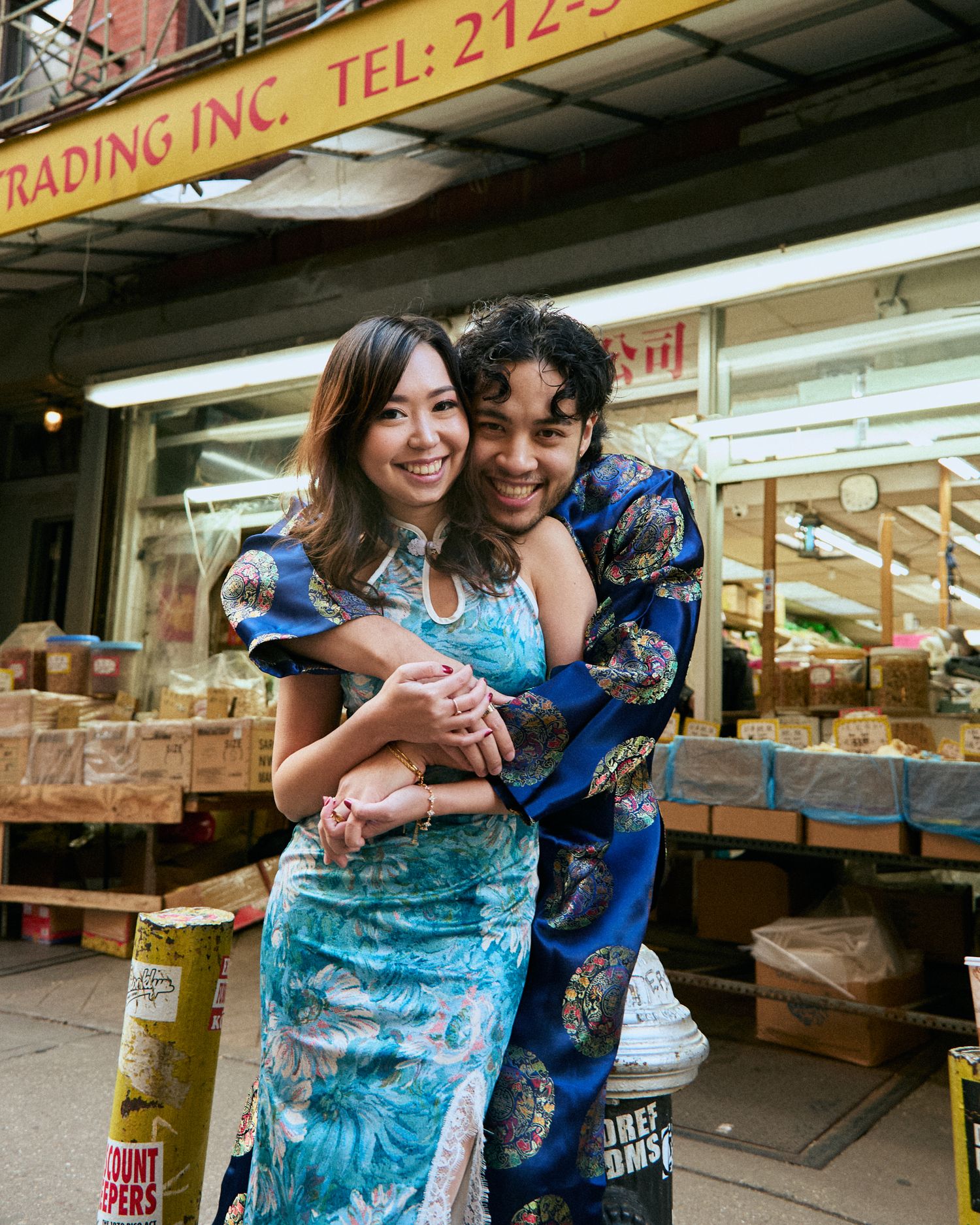 Couple hugging and smiling in front of a Chinatown storefront, wearing patterned outfits in daylight.