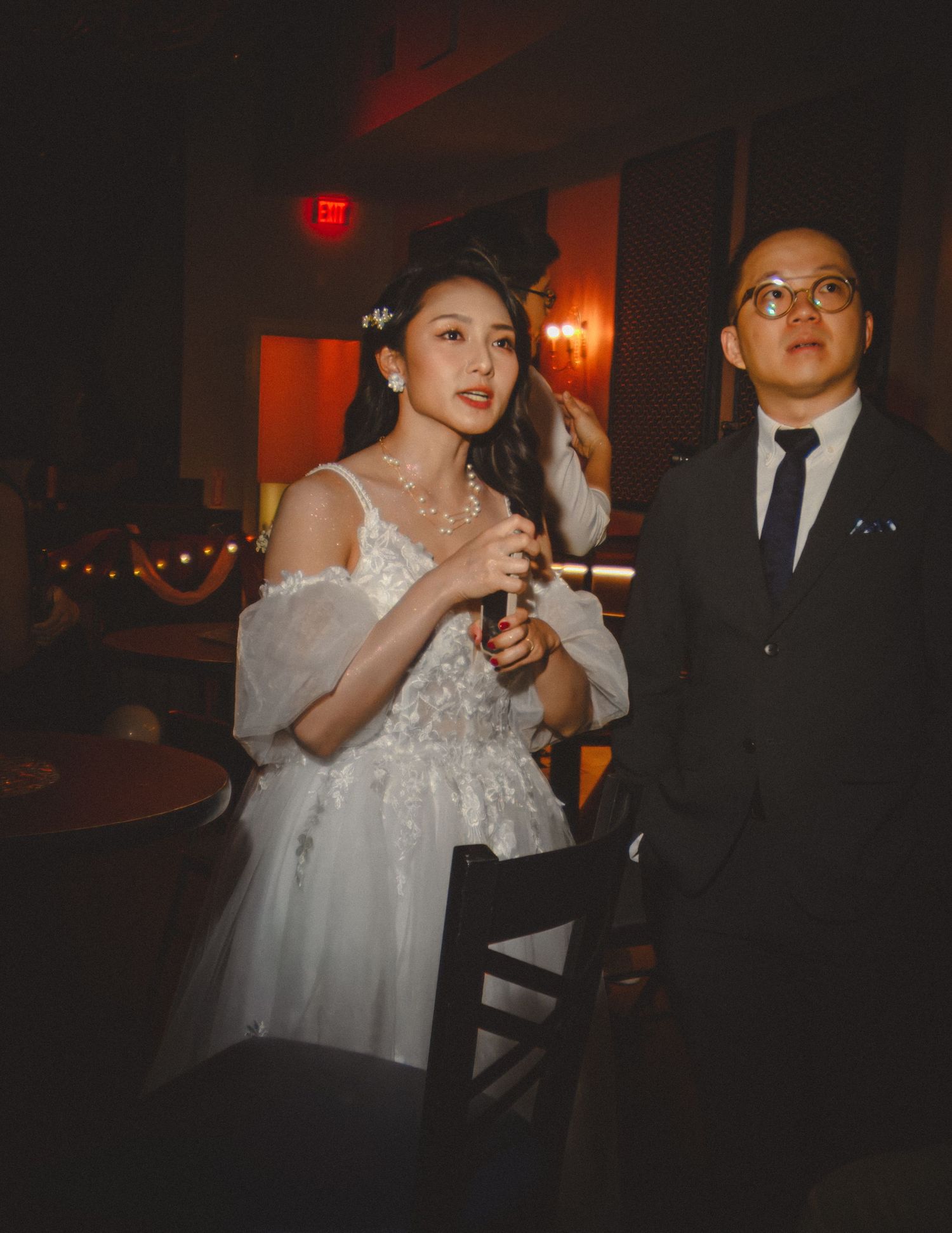 Bride in an off-shoulder white gown and groom in a dark suit posing indoors under warm, moody lighting.