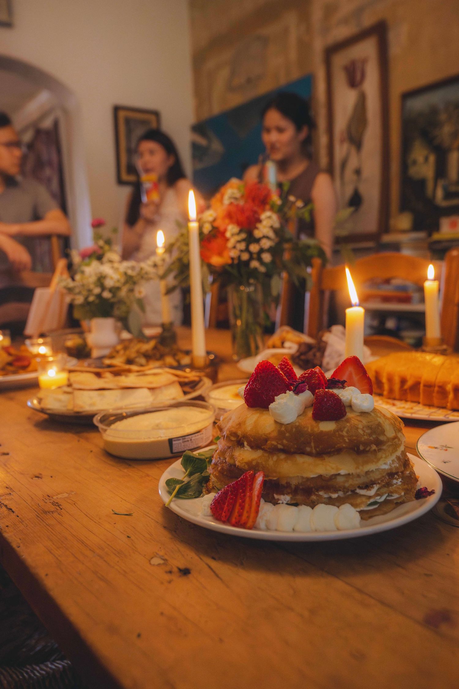 Candlelit dinner table with a strawberry-topped cake in the foreground and guests standing behind the table in a warmly lit room.