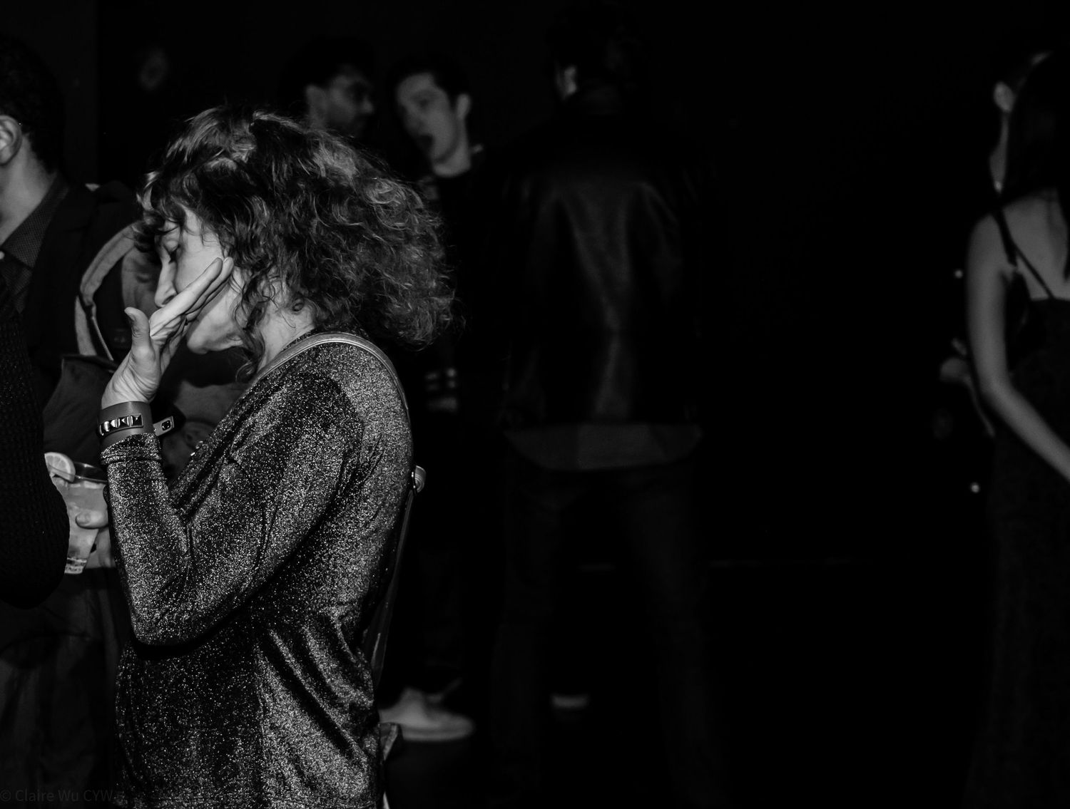 Black-and-white photo of a woman in a dim nightclub, turned in profile with her hand near her face while people blur in the background.