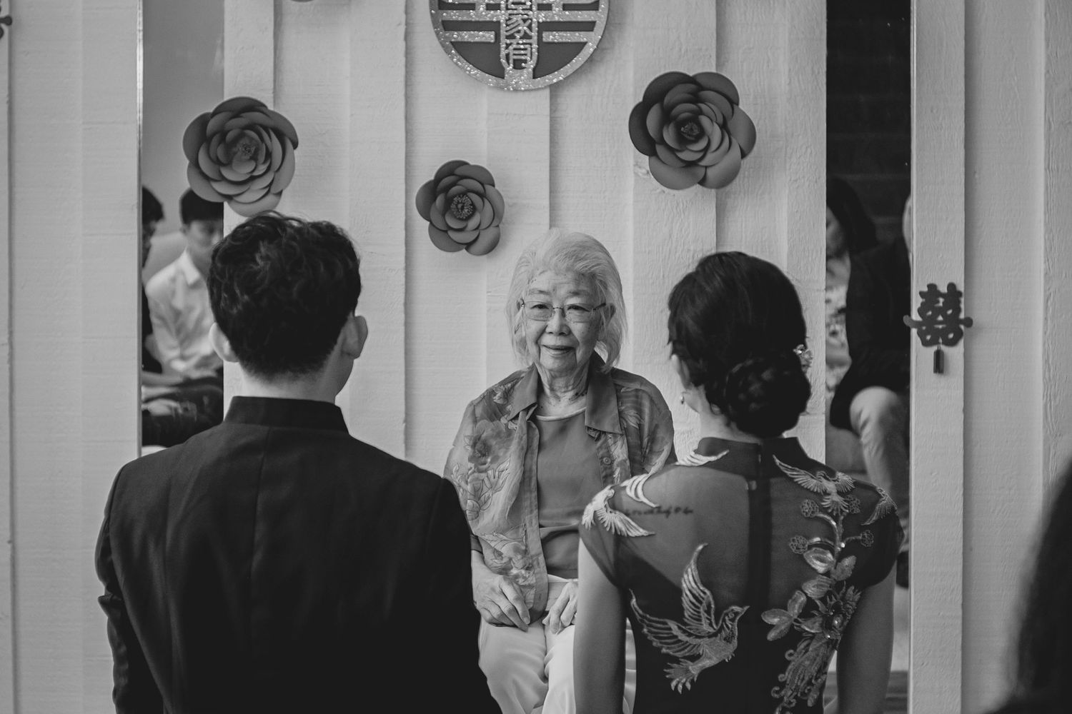 Black-and-white photo of an elderly woman smiling at a couple standing in a doorway decorated with large flower ornaments.