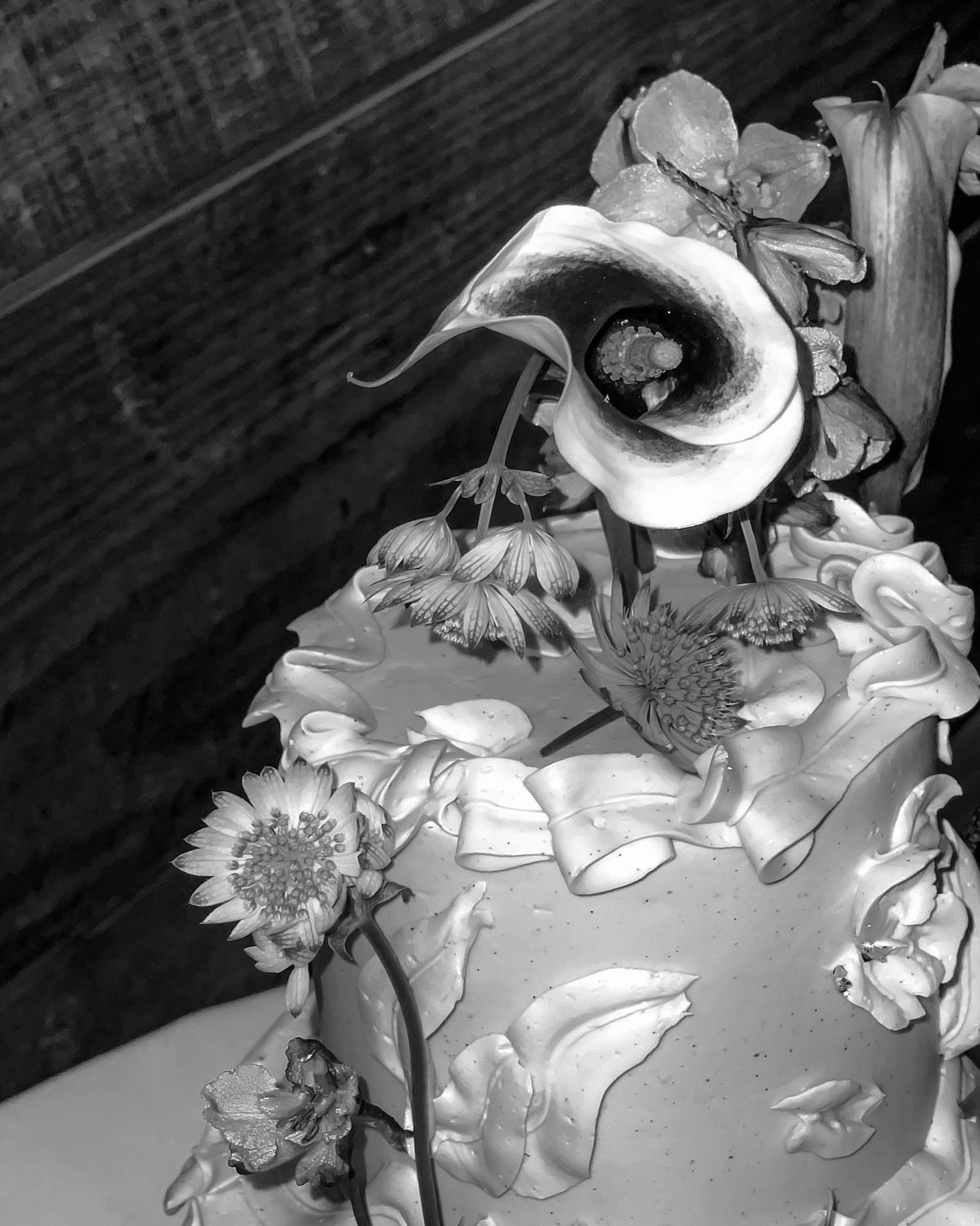 Black-and-white close-up of a decorated cake topped with flowers, including a calla lily and small blooms.
