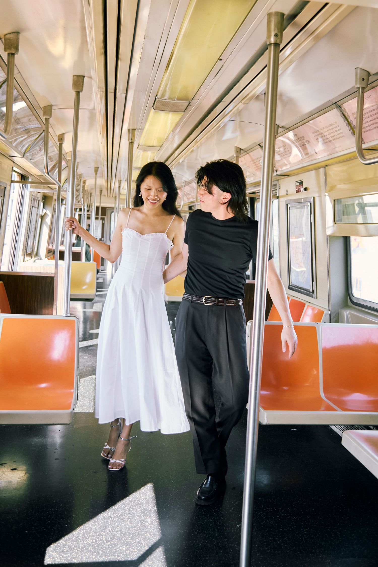 Couple standing in the middle of a subway car holding a pole, bright daylight filling the train with orange seats behind them.