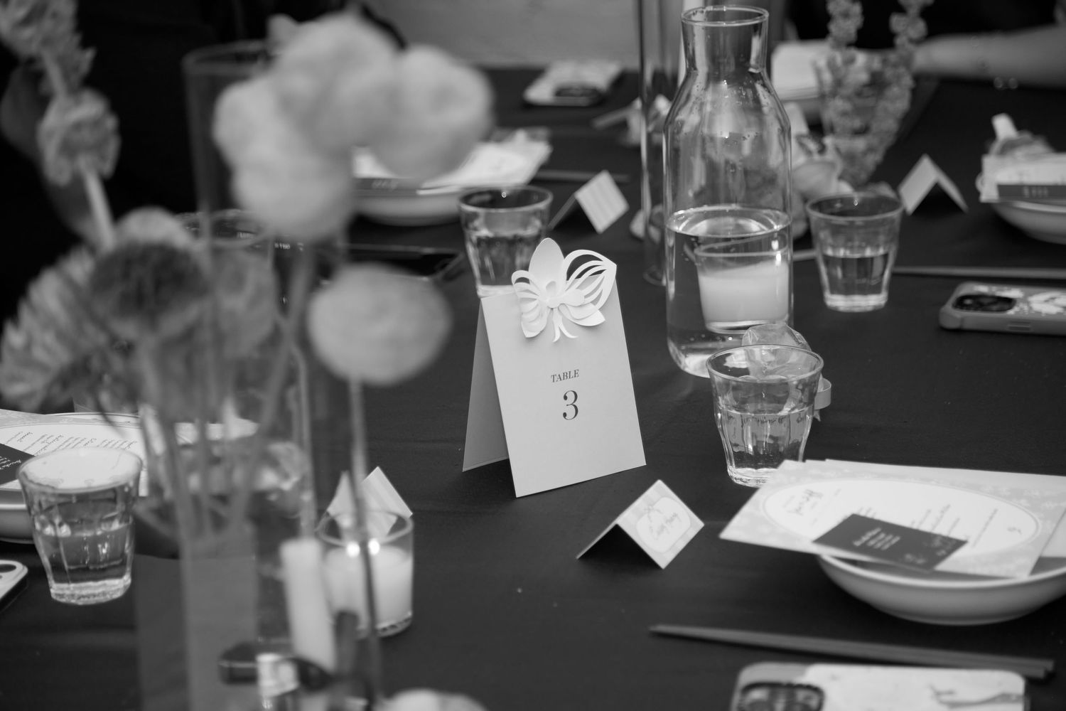 Black-and-white close-up of a dinner table with glasses and a folded table number card, with flowers blurred in the foreground.