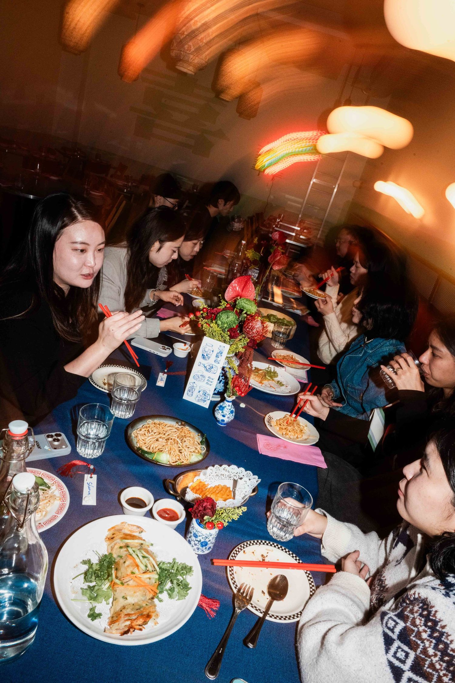 Flash photo of a group gathered around a long dinner table with shared dishes, with motion blur from warm lights overhead.