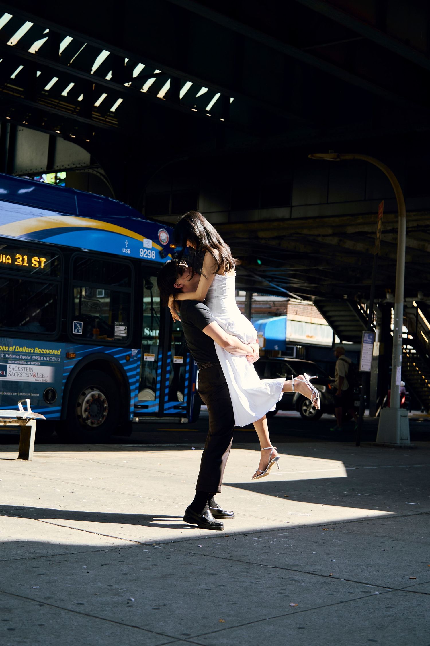 Couple kissing under an elevated structure as one partner lifts the other, with a city bus parked in the background.