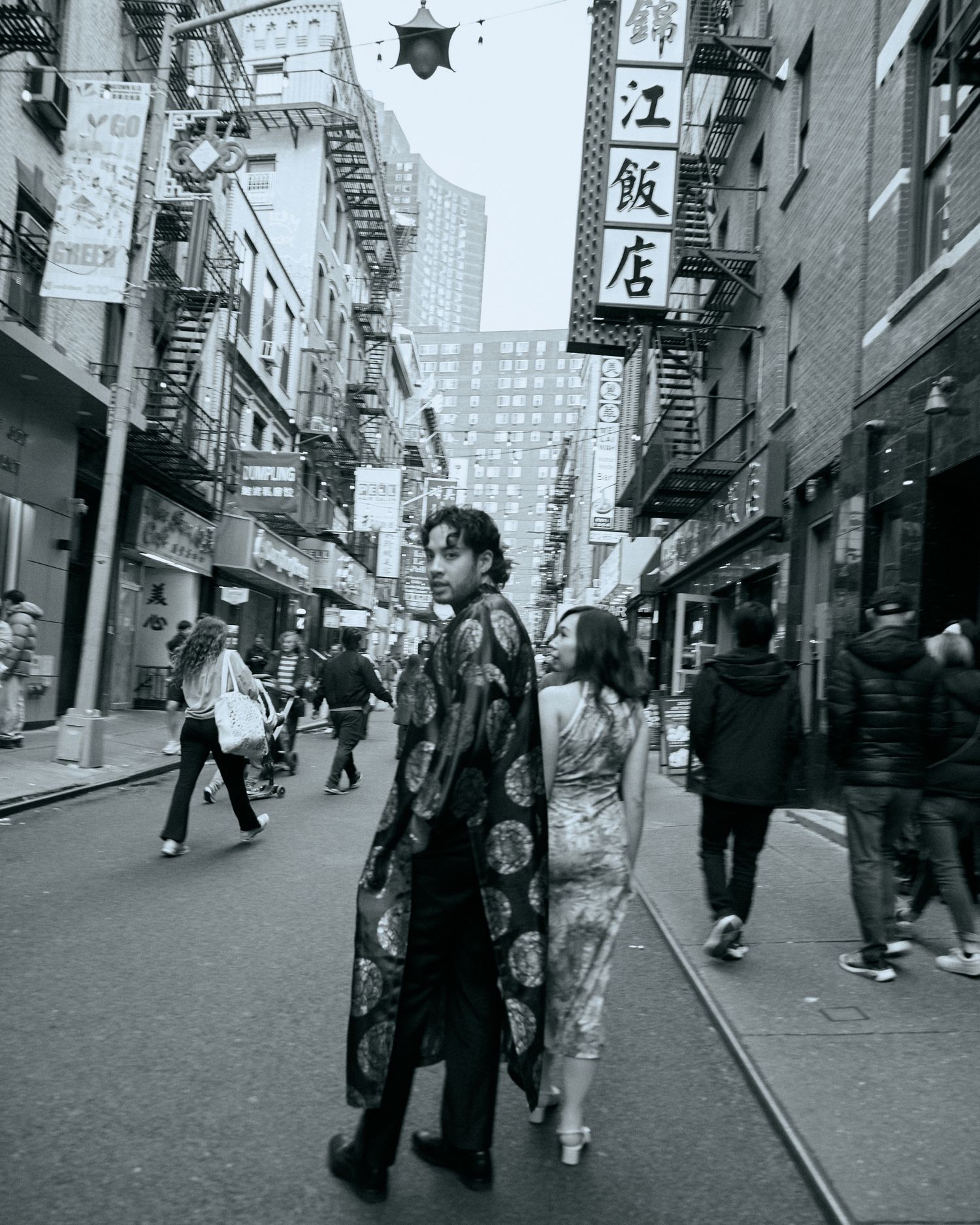 Black-and-white photo of a person standing in the middle of a Chinatown street, with storefront signs and pedestrians around.