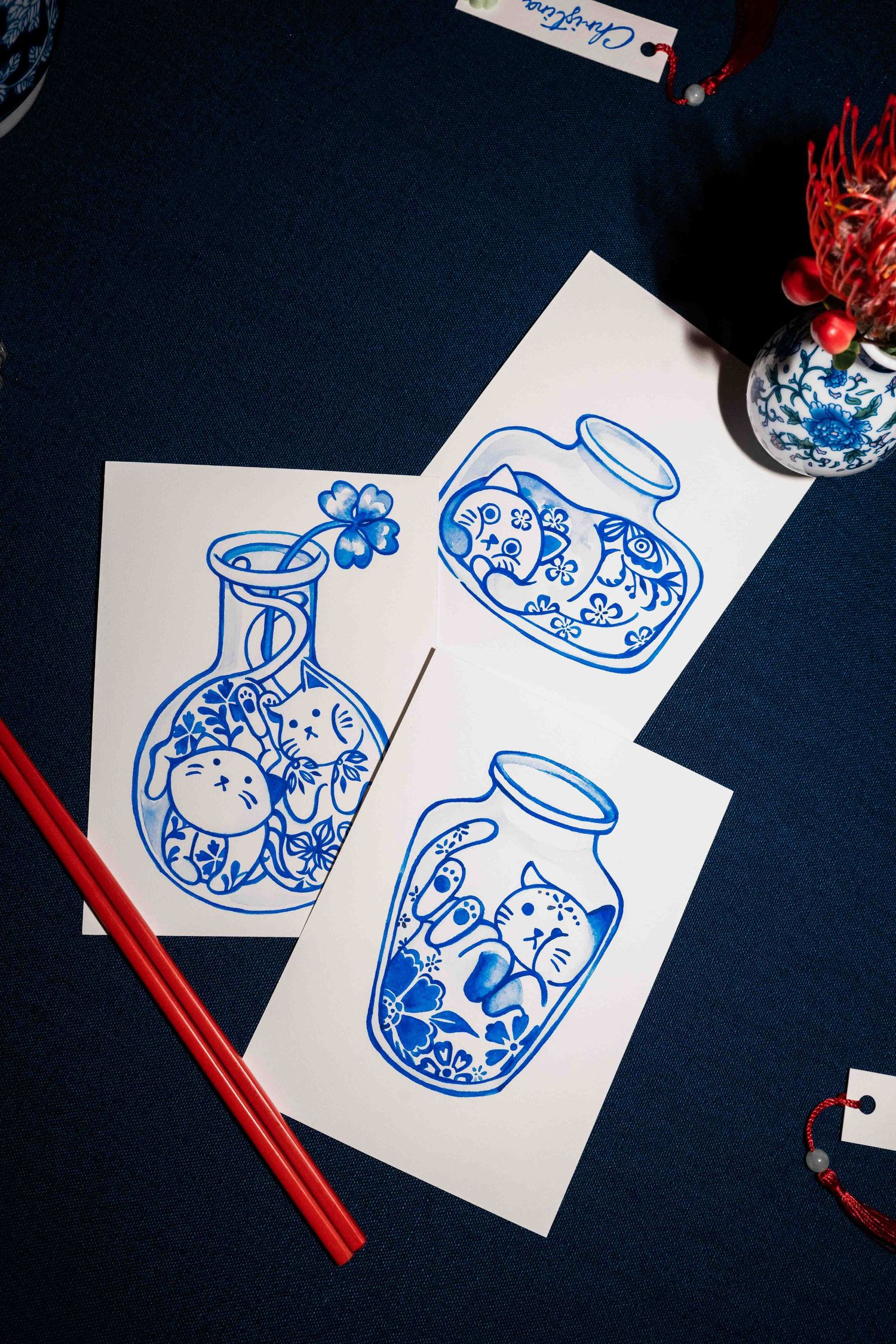Flat lay of blue-and-white illustrated cards featuring porcelain vase designs on a dark table, with red chopsticks placed beside them.