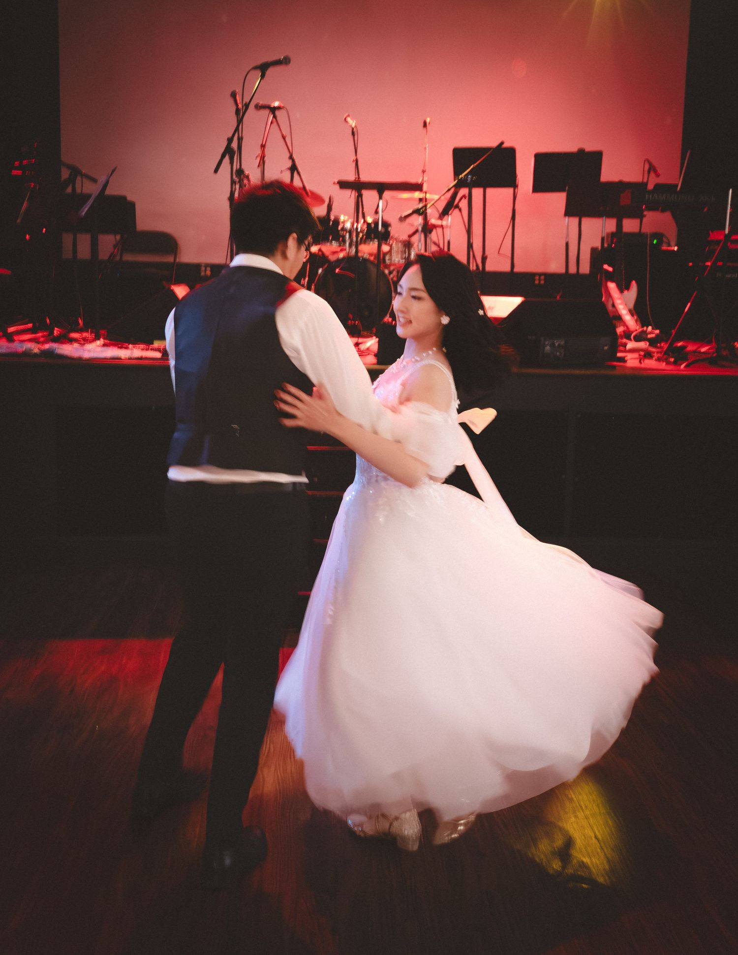 Couple dancing on a ballroom floor in front of a stage with instruments, lit by warm red lighting.