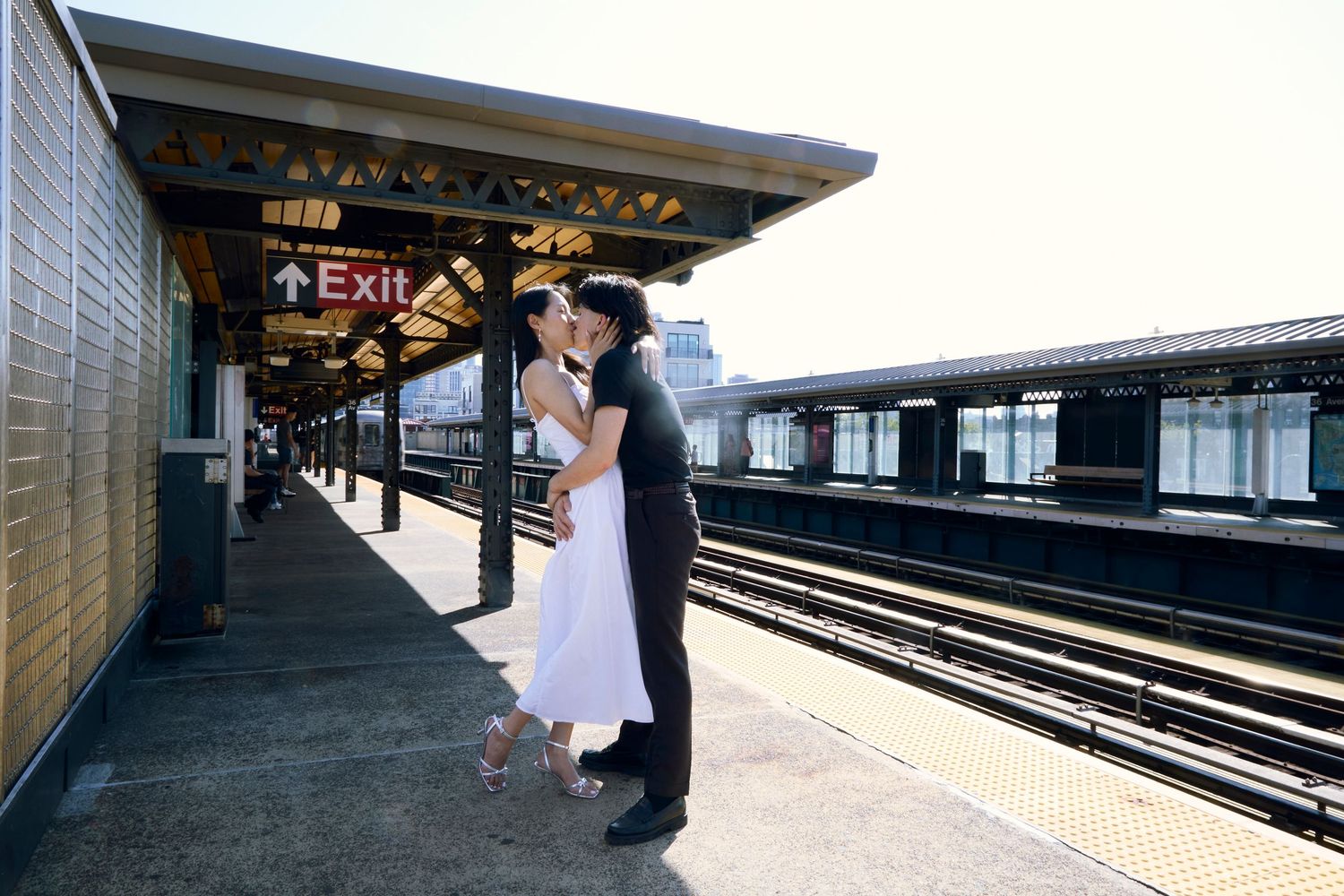 Couple embracing on a sunny subway platform beside the tracks, standing under the station canopy.