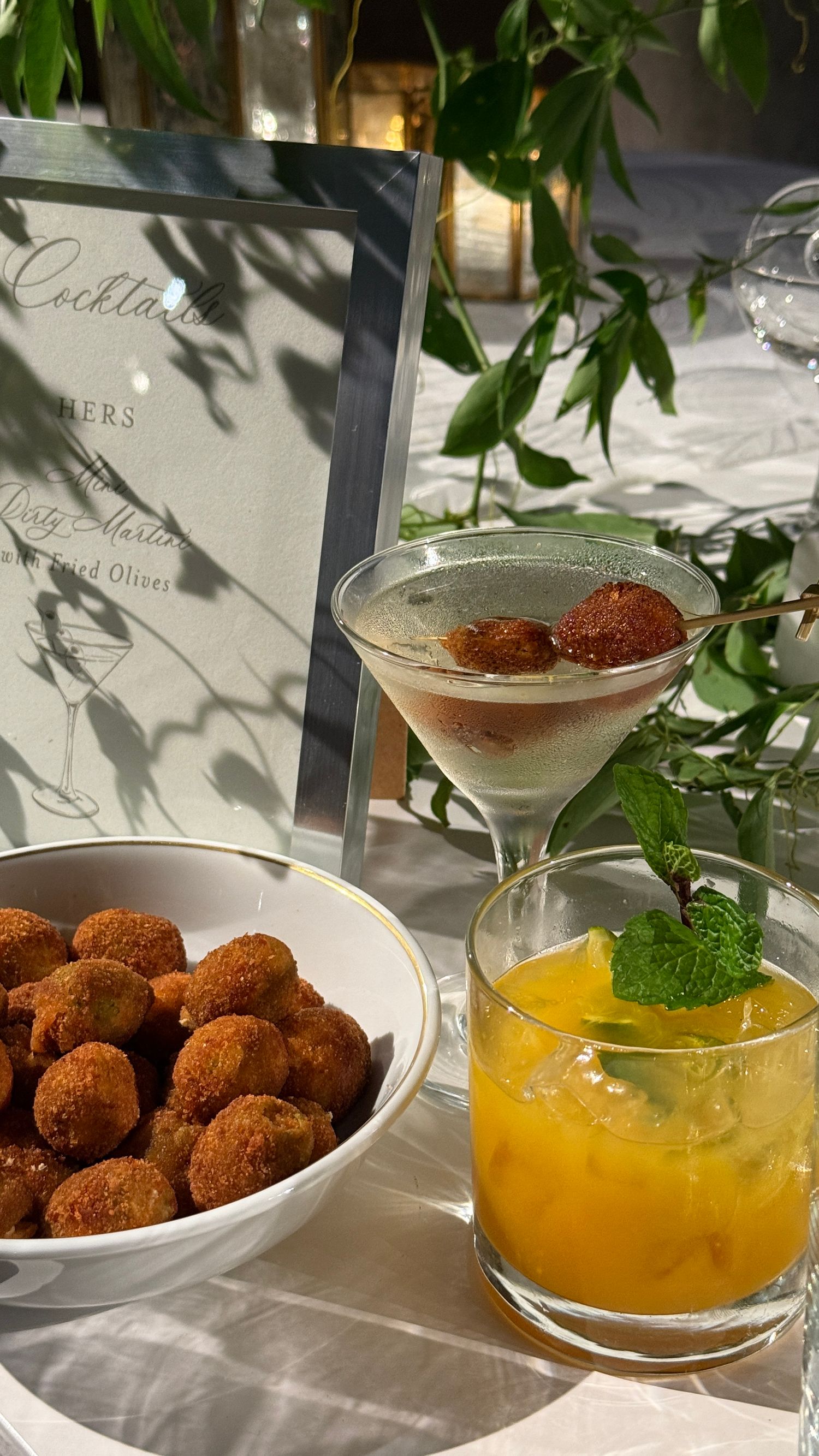 Sunlit table with cocktails, a bowl of snacks, and leafy greenery casting shadows beside a printed menu.