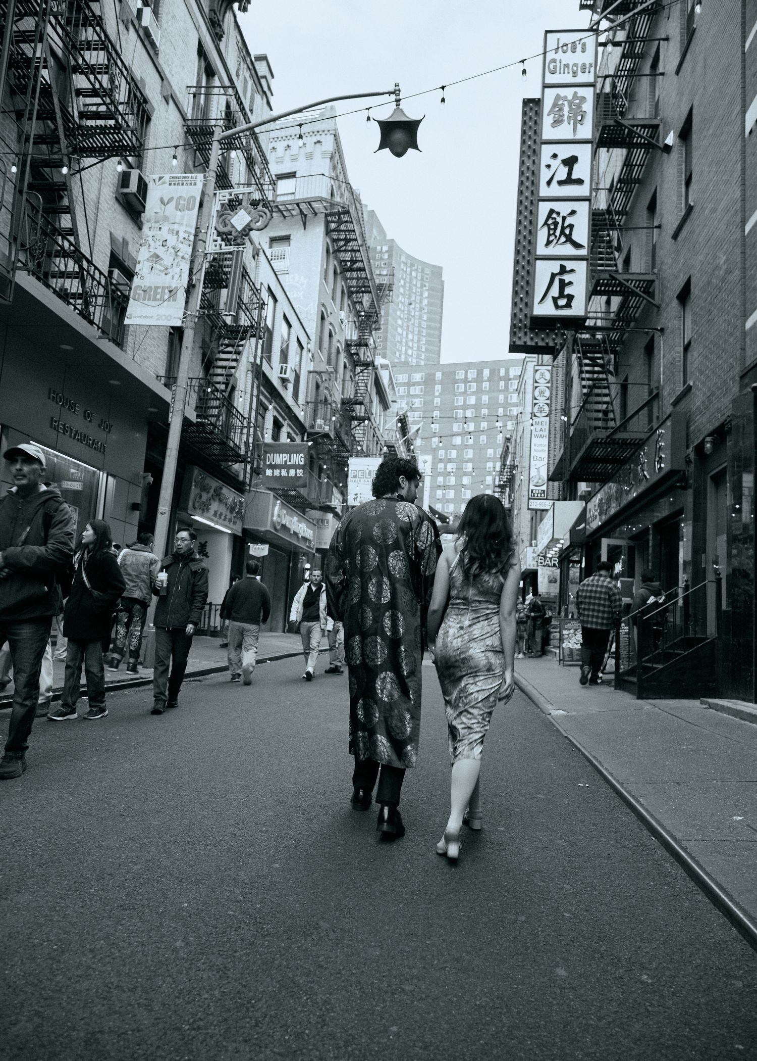 Black-and-white photo of a couple walking down a Chinatown street, surrounded by storefront signs and pedestrians.