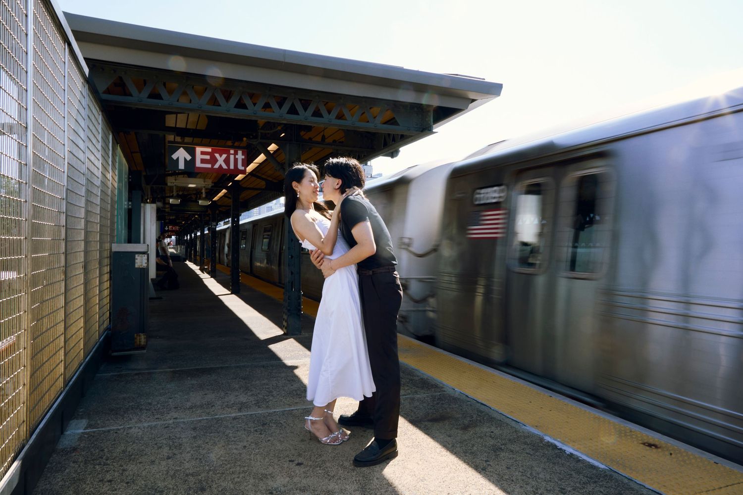 Couple kissing on an outdoor subway platform as a train passes by, with an exit sign above the platform.