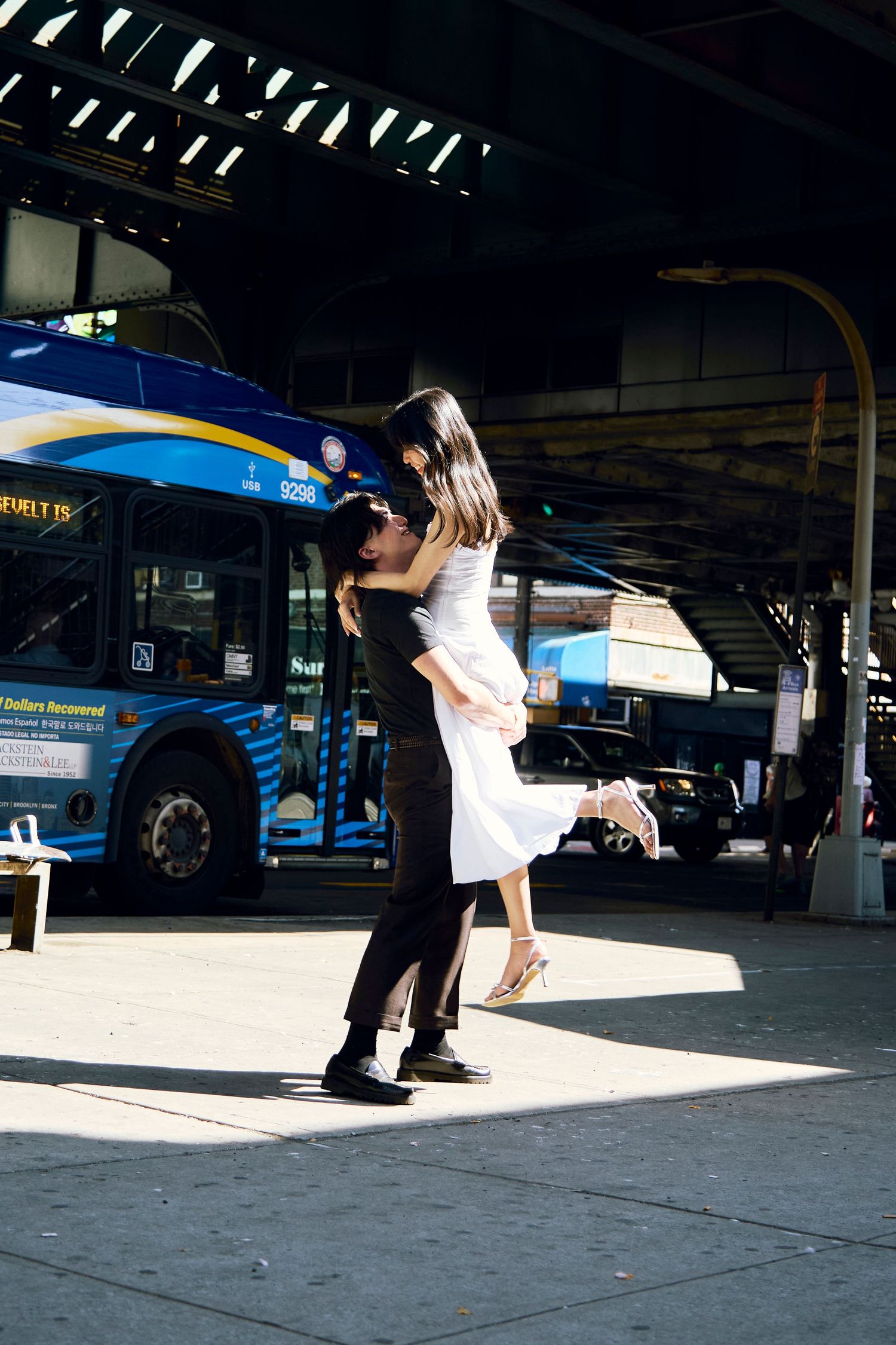 Couple embracing and kissing in bright sunlight on a sidewalk near a blue bus under an overpass.