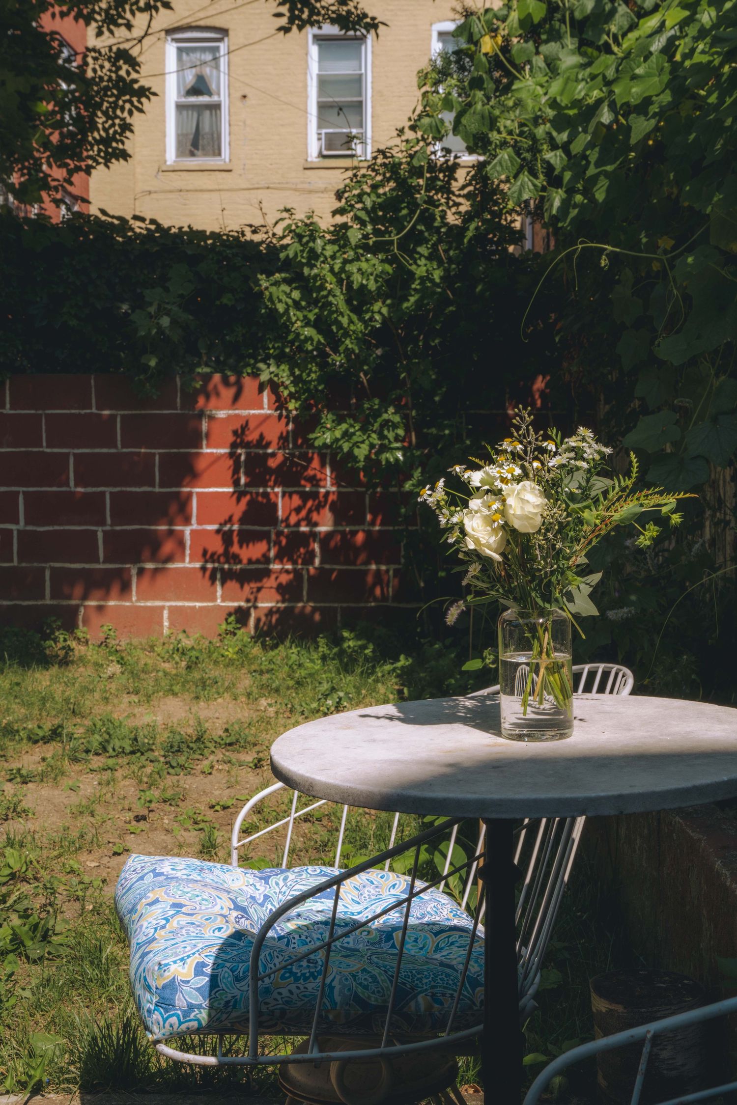 Sunlit backyard corner with a small round metal table, patterned chair cushions, and a vase of flowers on the tabletop.