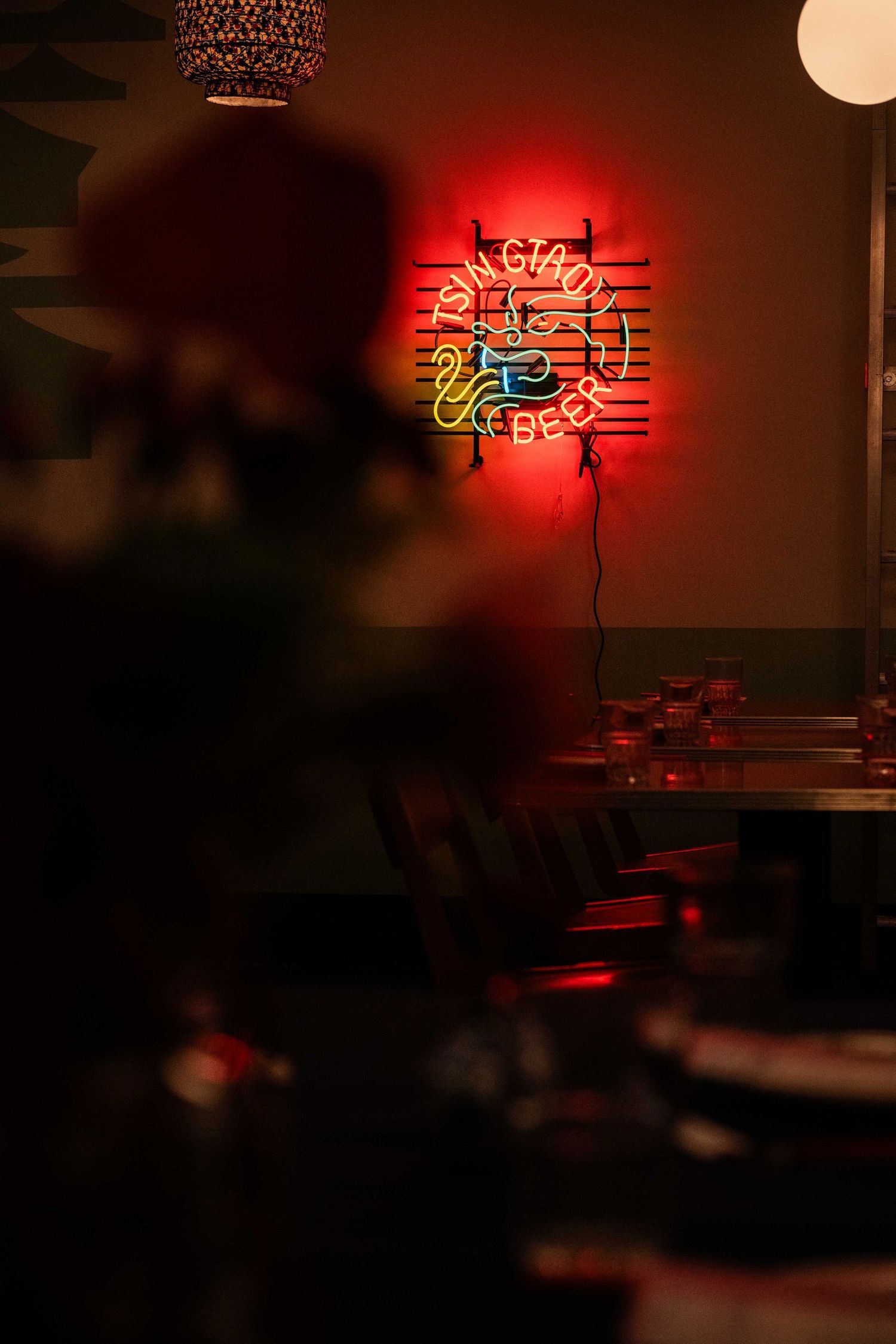 Moody indoor photo of a glowing red neon sign on a wall, with tables and chairs in soft focus in the foreground.