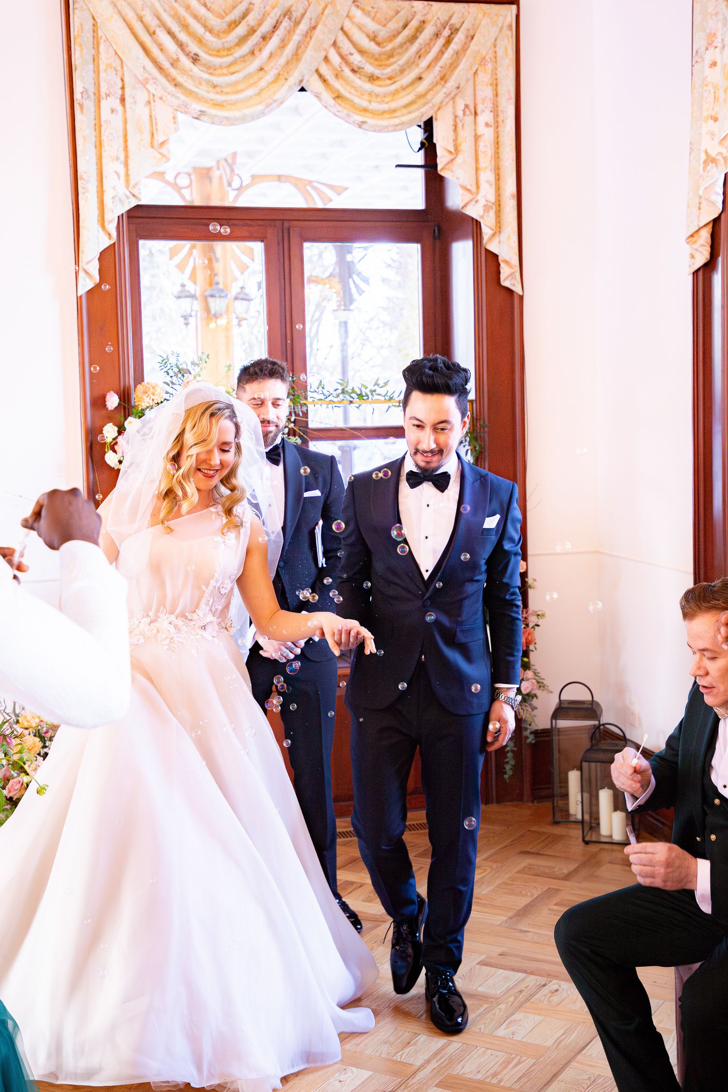 Wedding party celebrating in an elegant ballroom with white decor and wooden floors during a joyful dance moment.