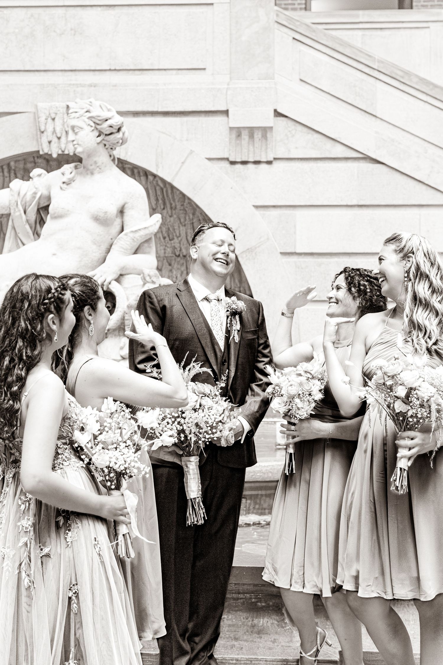 Bridesmaids in champagne-colored dresses share a candid laughing moment while holding bouquets at an outdoor wedding.