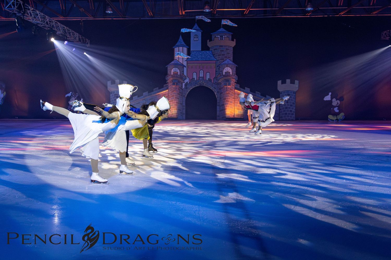 Disney on Ice performers in character costumes skating on ice rink with colorful stage lighting and castle backdrop.