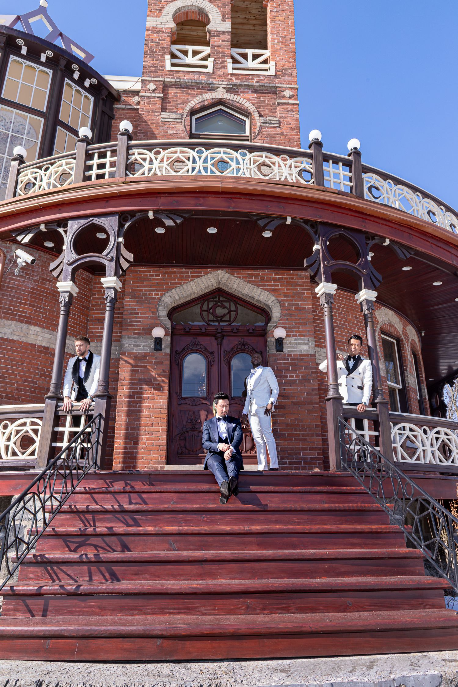 Wedding party poses on red wooden steps in front of historic brick building with ornate white railings.